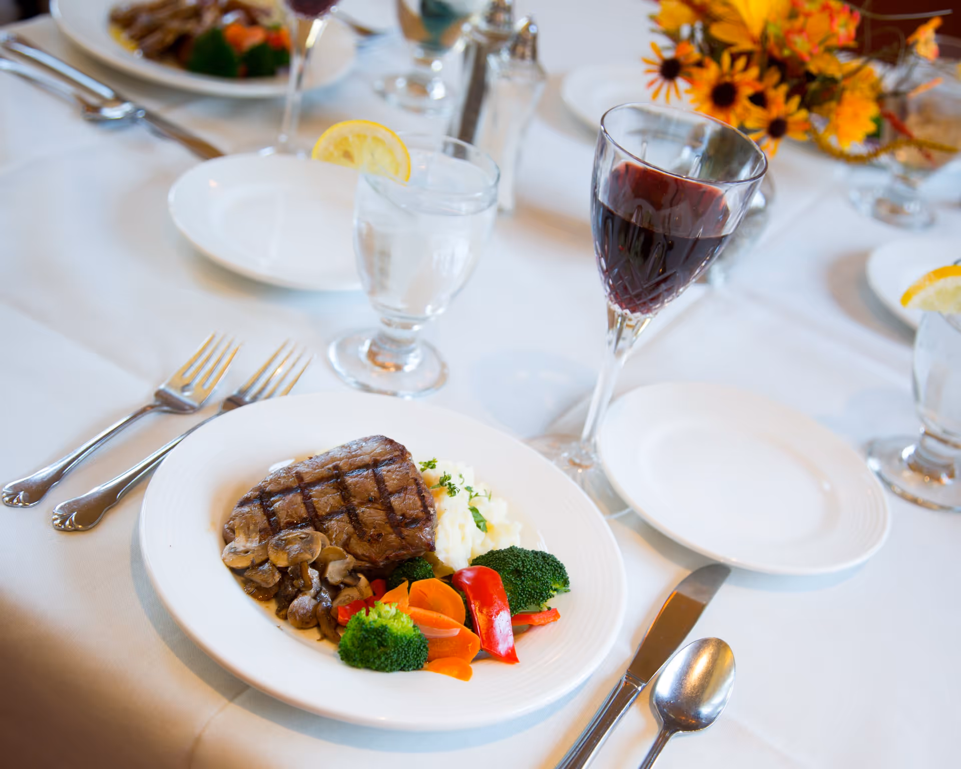 A plated meal on a white tablecloth featuring a grilled steak with mushrooms, mashed potatoes, broccoli, carrots, and red bell peppers. Next to the plate is a glass of red wine and a glass of water with a lemon slice. The table is set with silverware and additional plates, with a floral centerpiece in the background.