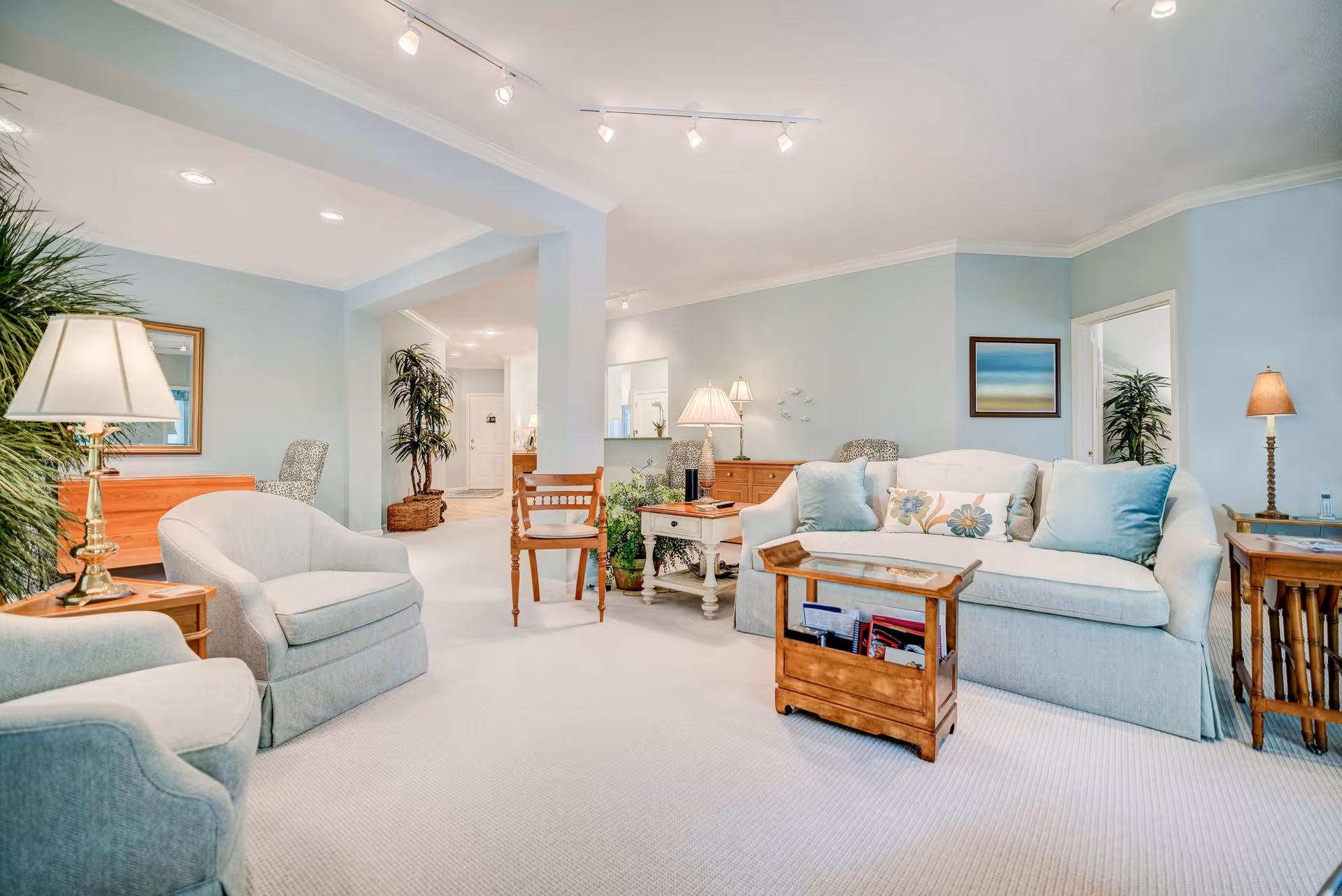 Bright, spacious living room with light blue walls, upholstered sofa and armchairs, wooden coffee and side tables, lamps, and potted plants.