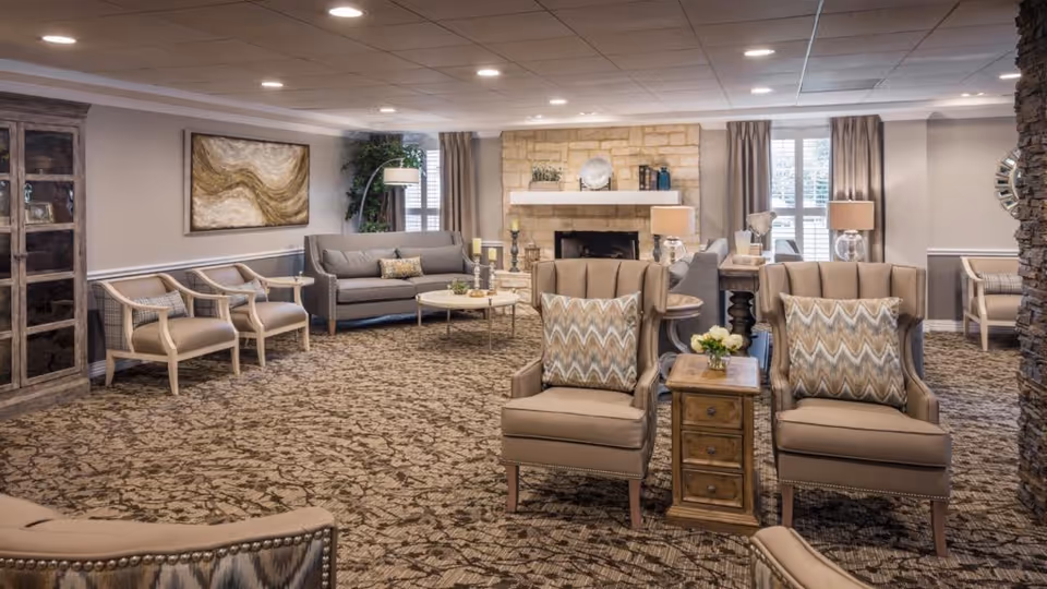 A spacious senior living common area with patterned carpet, two beige armchairs with decorative pillows, a wooden side table with a small flower arrangement, a gray sofa, several chairs, a stone fireplace, and large windows with curtains allowing natural light.