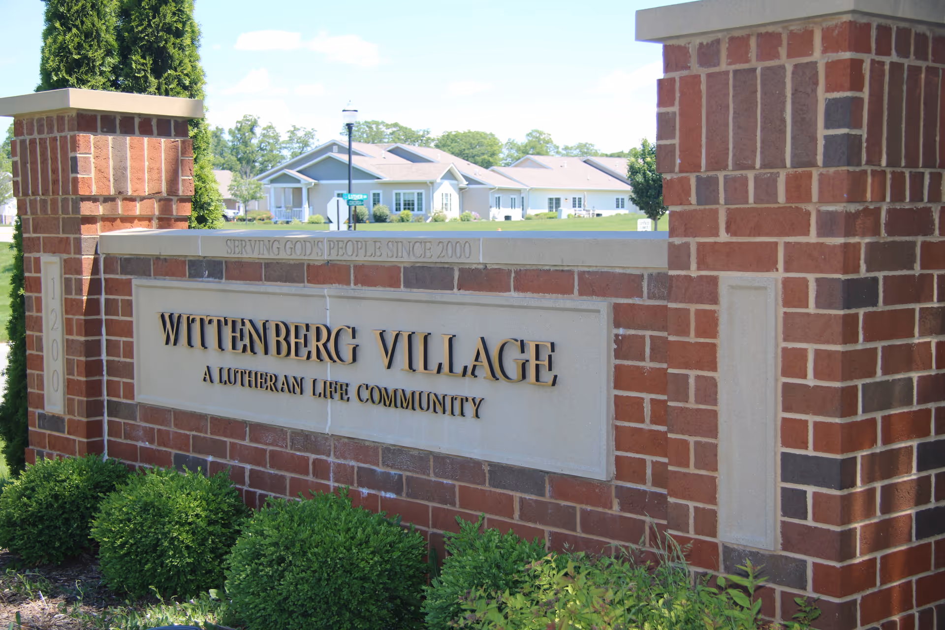Brick entrance sign for Wittenberg Village, a Lutheran Life Community, with neatly trimmed bushes in front and residential houses visible in the background under a clear sky.