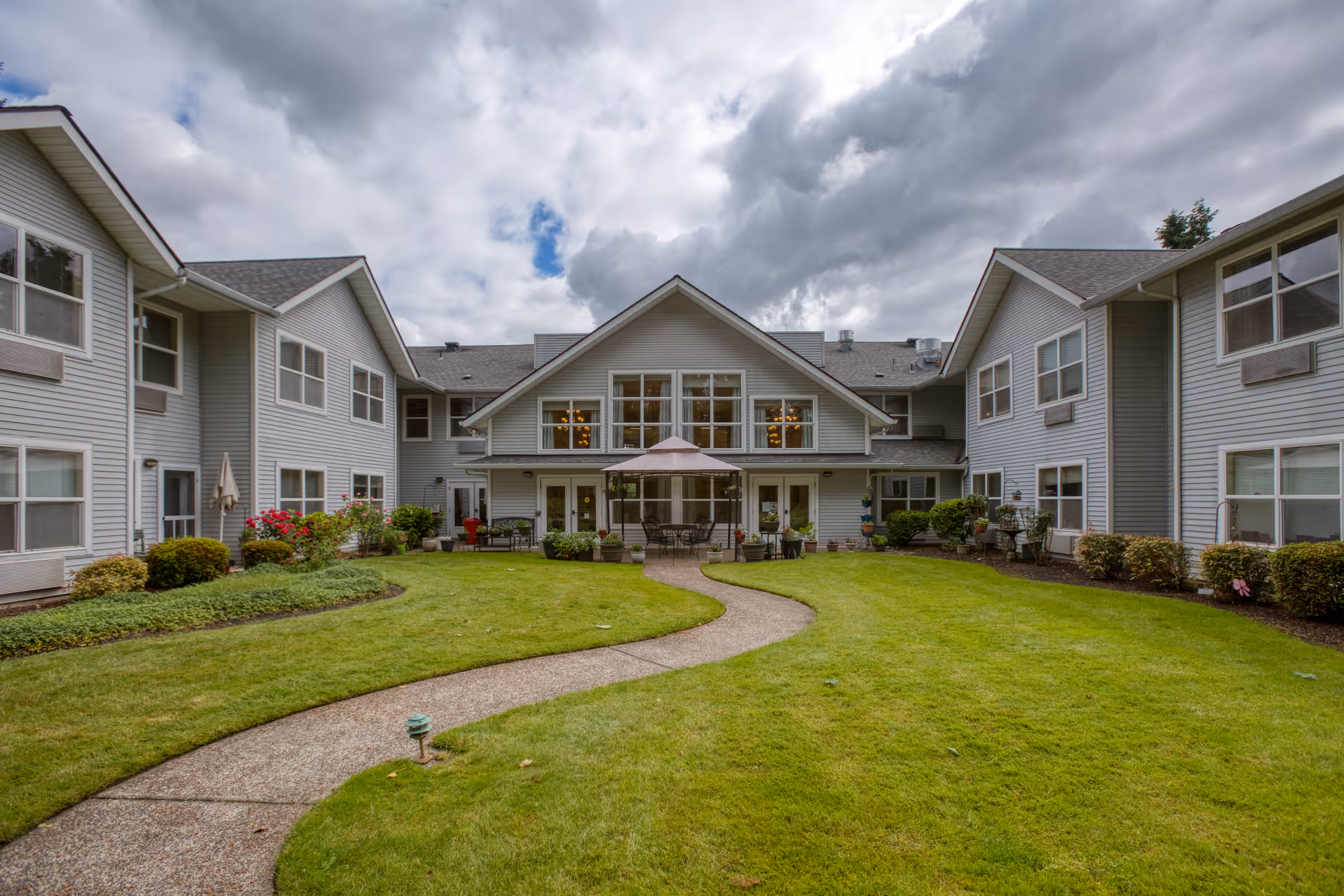 View of a two-story assisted living facility building with gray siding surrounding a well-maintained green lawn and a curved concrete pathway leading to a patio area with tables, chairs, and an umbrella. The sky is partly cloudy.