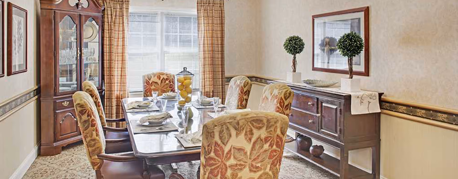 Formal dining room with a set table, upholstered floral chairs, a china cabinet and a sideboard beneath a window.