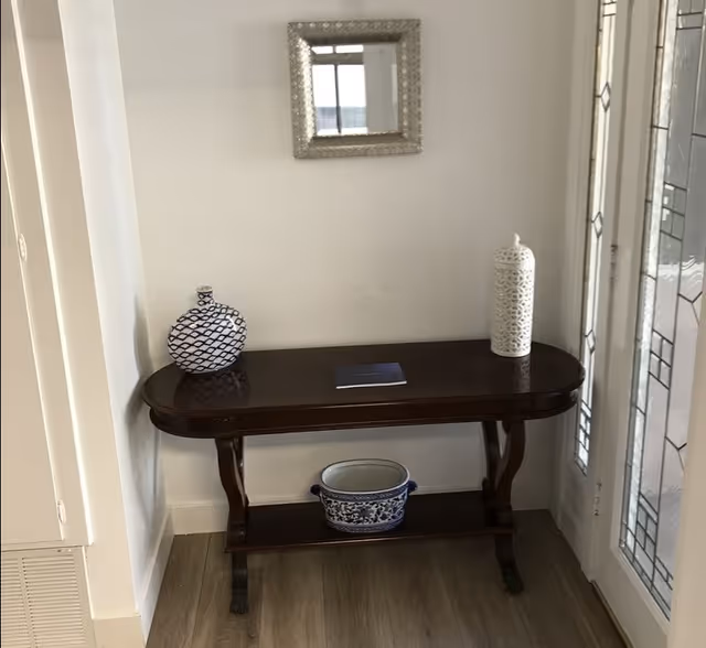A small wooden console table with curved legs placed against a white wall. On top of the table are two decorative vases, one white with a lattice pattern and the other blue and white with a geometric pattern. A small rectangular mirror with a textured frame hangs on the wall above the table. Below the table is a blue and white ceramic pot. To the right of the table is a door with decorative glass panels.