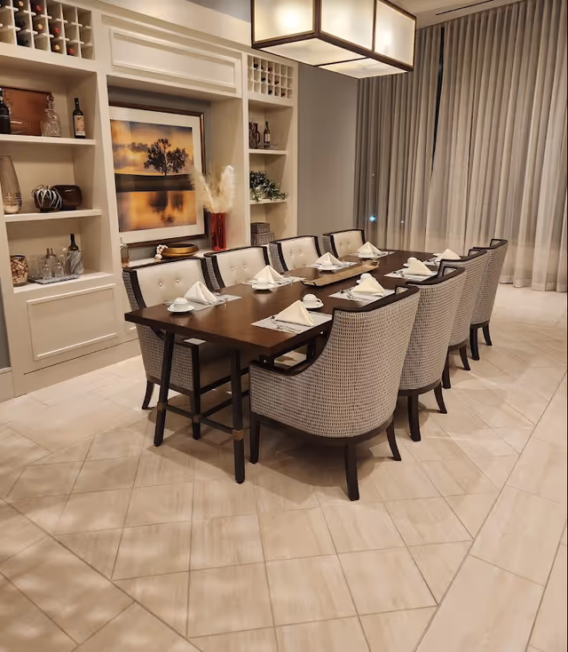 A formal dining room featuring a long wooden table set with napkins and cups, surrounded by upholstered chairs and built-in shelving.
