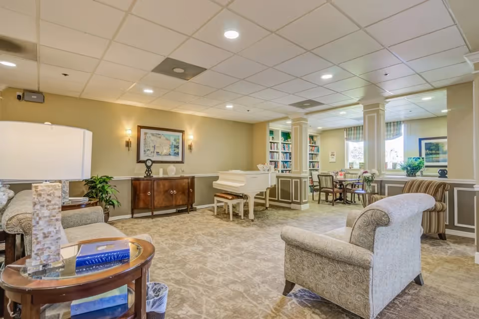 Community lounge with upholstered armchairs, a white piano, bookshelves and a small dining table under recessed lighting.