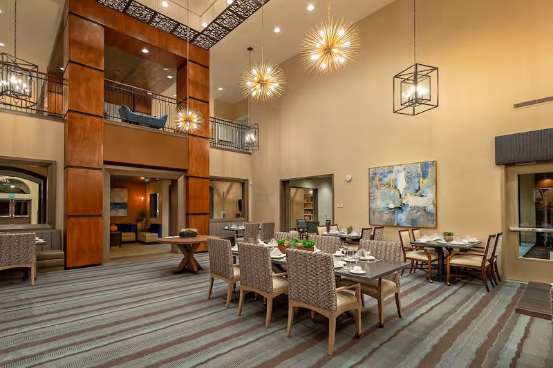 A spacious dining area in a senior living facility with multiple tables and chairs arranged neatly. The room features high ceilings with modern pendant lighting fixtures, a large abstract painting on the wall, and a carpeted floor with striped patterns. There is a balcony area above and an adjacent lounge area visible through an open doorway.