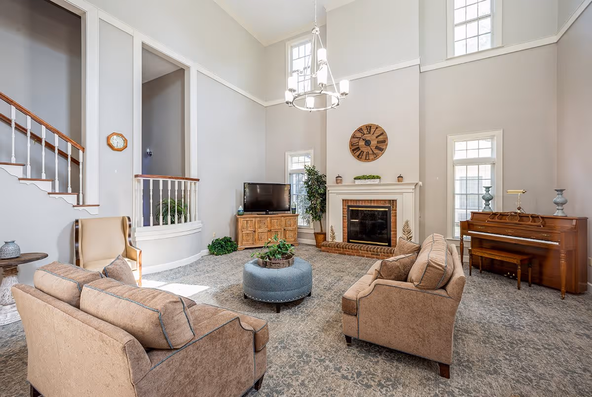 A spacious living room with high ceilings and large windows allowing natural light to fill the space. The room features two beige sofas facing each other with a round blue ottoman in the center. There is a wooden piano against the right wall, a fireplace with a large clock above it, a TV on a wooden stand, and a staircase with white railings on the left side. The walls are painted light gray and the floor is covered with a patterned carpet.