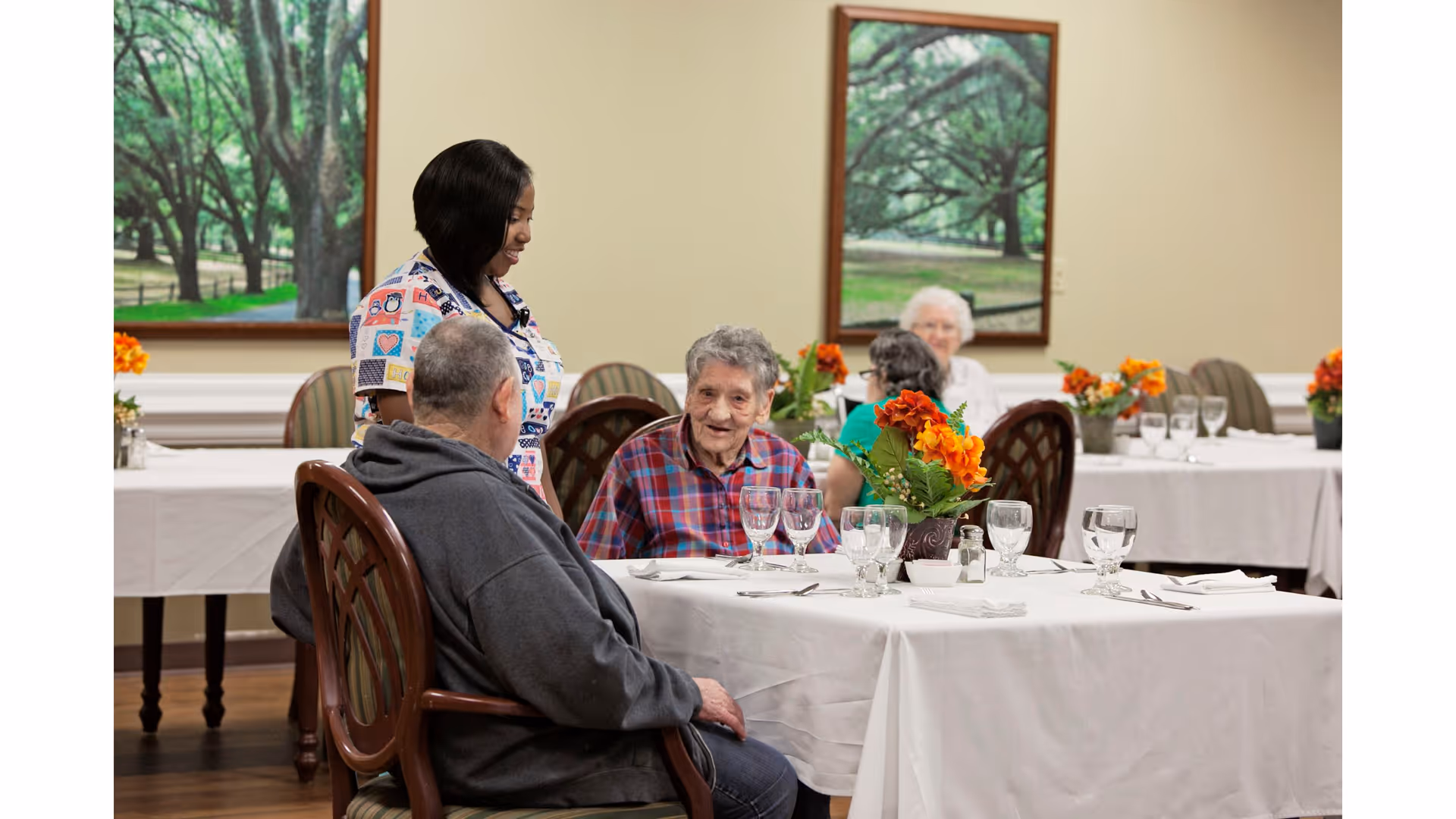 A caregiver stands while residents sit at a table set with glassware and orange floral centerpieces in a communal dining room.