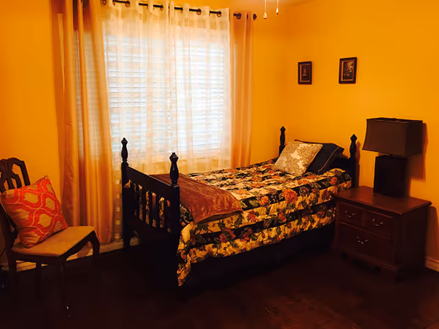 A cozy bedroom with a single bed covered in a floral quilt and a brown throw blanket. The bed has a decorative pillow and is positioned against a yellow wall. Next to the bed is a wooden nightstand with a dark-colored lamp. A wooden chair with a cushion and a decorative pillow is placed near a window with sheer white curtains and beige drapes. Two small framed pictures hang on the wall above the nightstand.