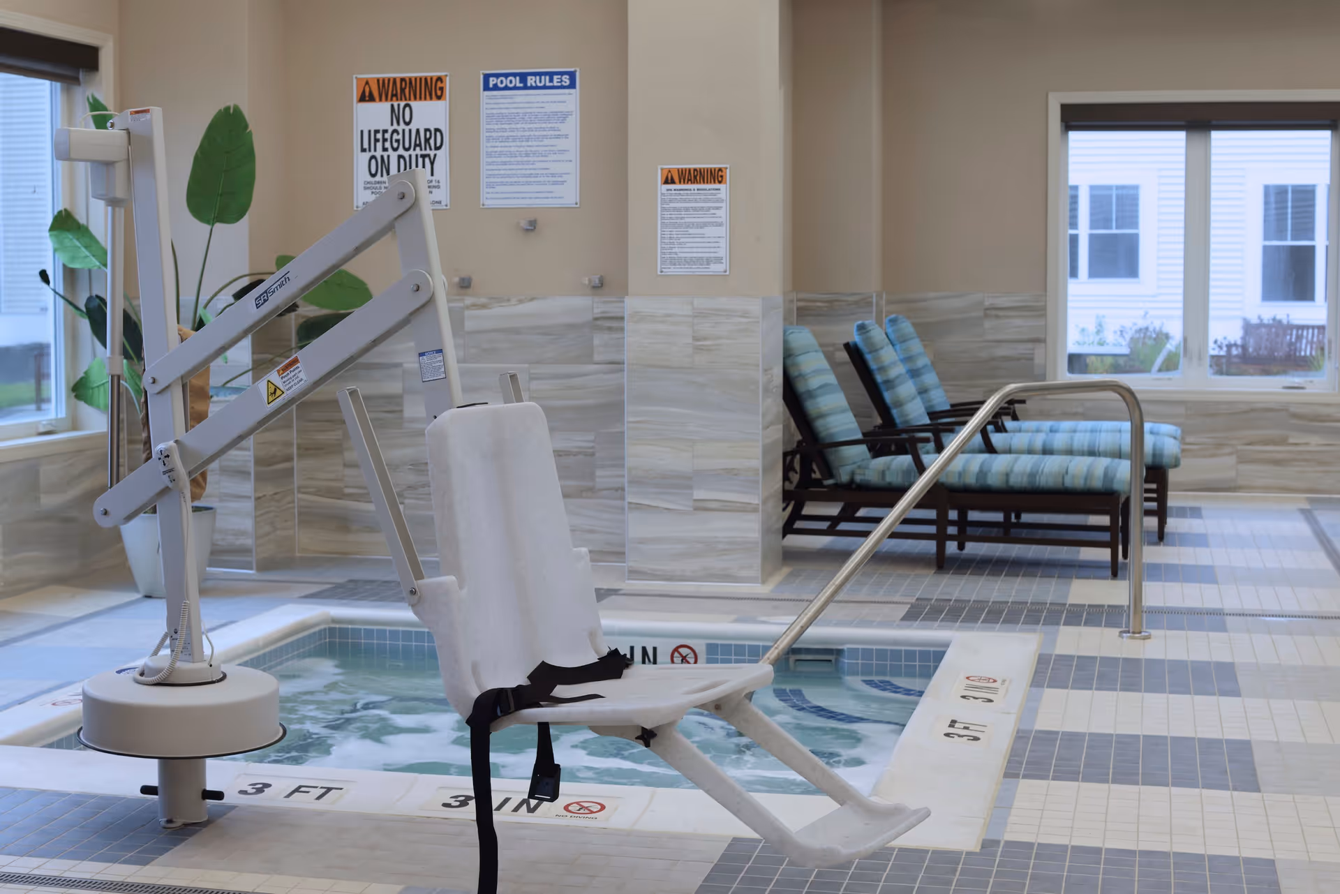 Indoor pool area with a white pool lift chair for accessibility, a tiled floor, and three cushioned lounge chairs with blue striped cushions near a window. Warning and pool rules signs are posted on the wall.