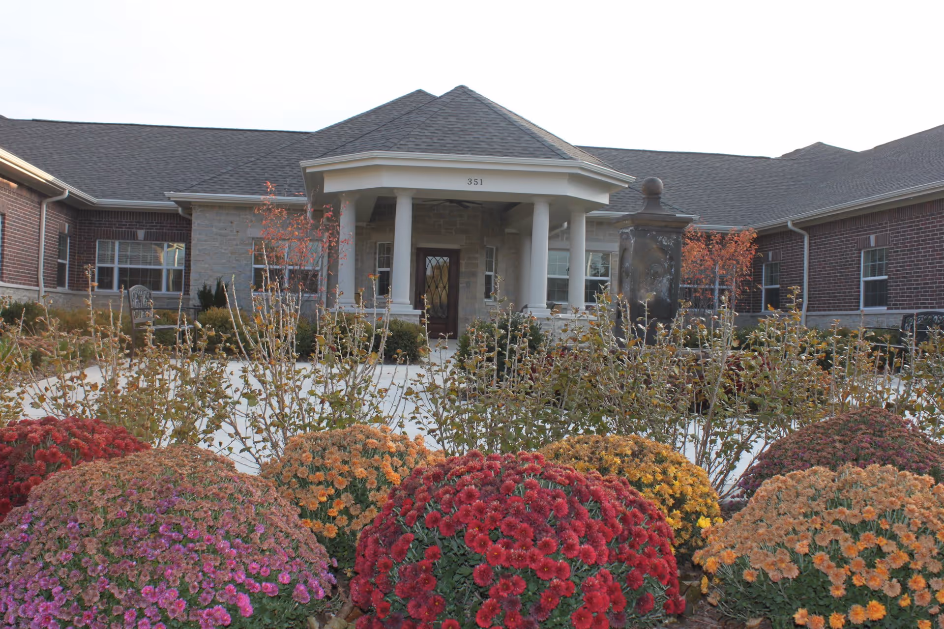 Front exterior view of a single-story brick and stone building with a covered entrance supported by white columns. The building is surrounded by a garden with colorful flowering bushes in shades of red, orange, yellow, and purple.