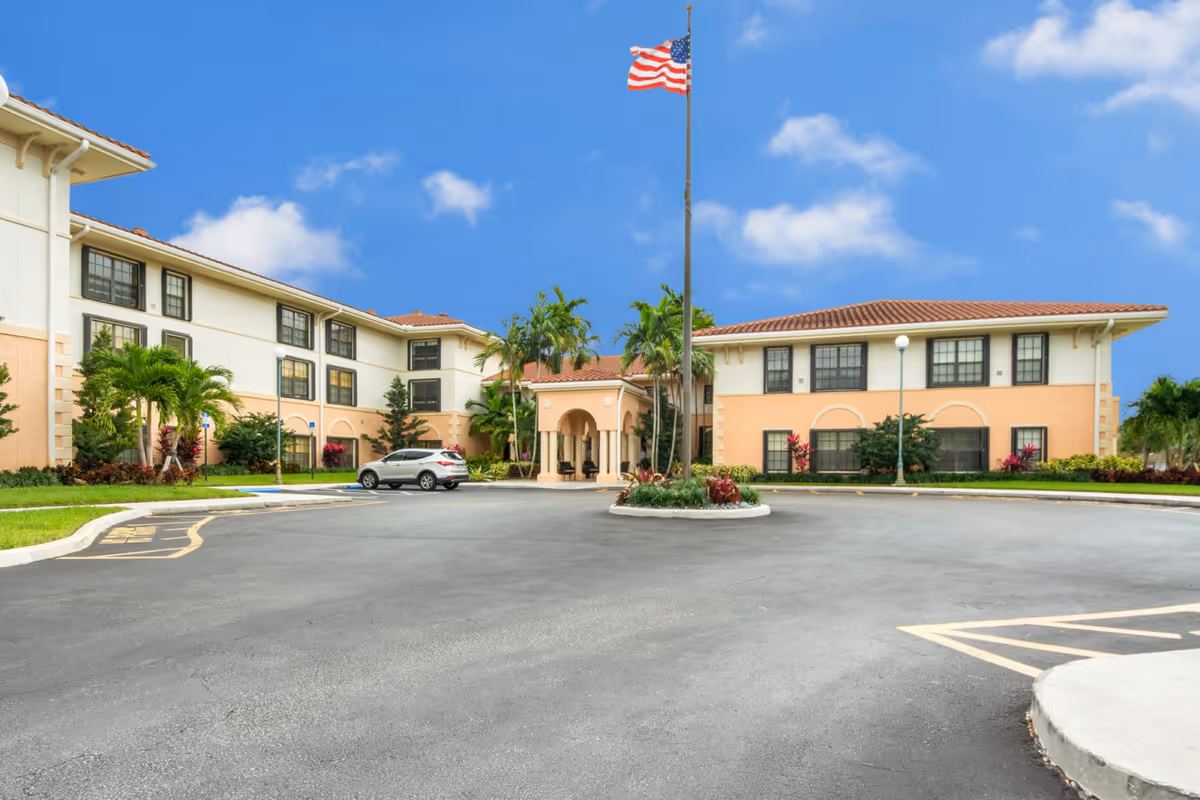 Exterior view of Presidential Place senior living facility showing a two-story building with beige and peach walls, black-framed windows, a circular driveway with a flagpole flying the American flag in the center, and landscaped greenery including palm trees and shrubs under a blue sky with scattered clouds.