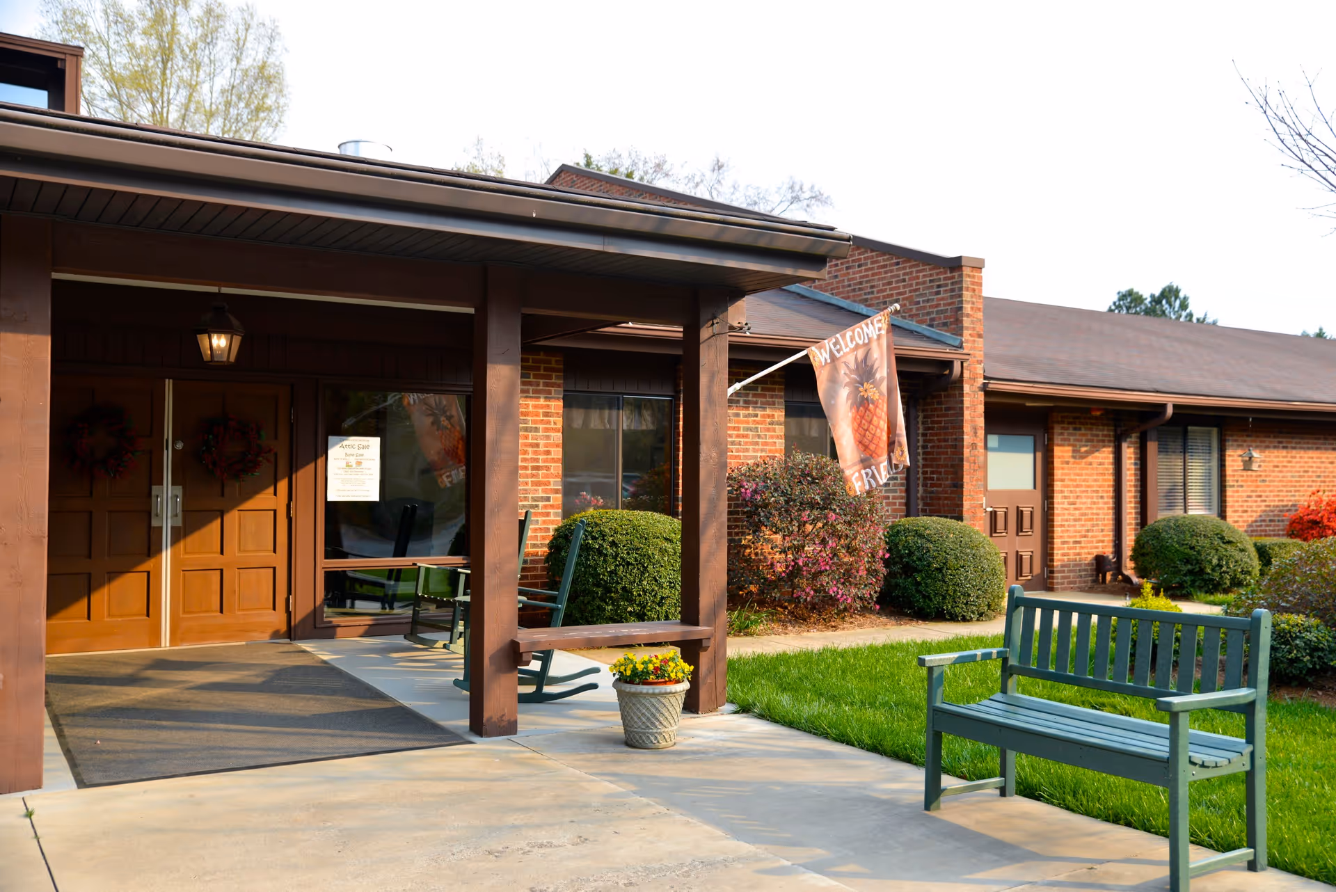 Entrance of a brick building with a covered porch featuring wooden double doors, two rocking chairs, a welcome flag with a pineapple design, a green bench on the sidewalk, and neatly trimmed bushes and grass surrounding the area.