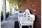 White wooden benches on a covered brick-front porch with columns and a concrete walkway.