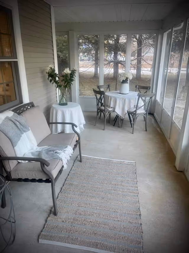 A screened-in porch with a cushioned bench covered with a gray and white throw blanket, a small round table with a white tablecloth and a vase of flowers, and a round dining table with a white tablecloth and four chairs. The porch overlooks a yard with trees.