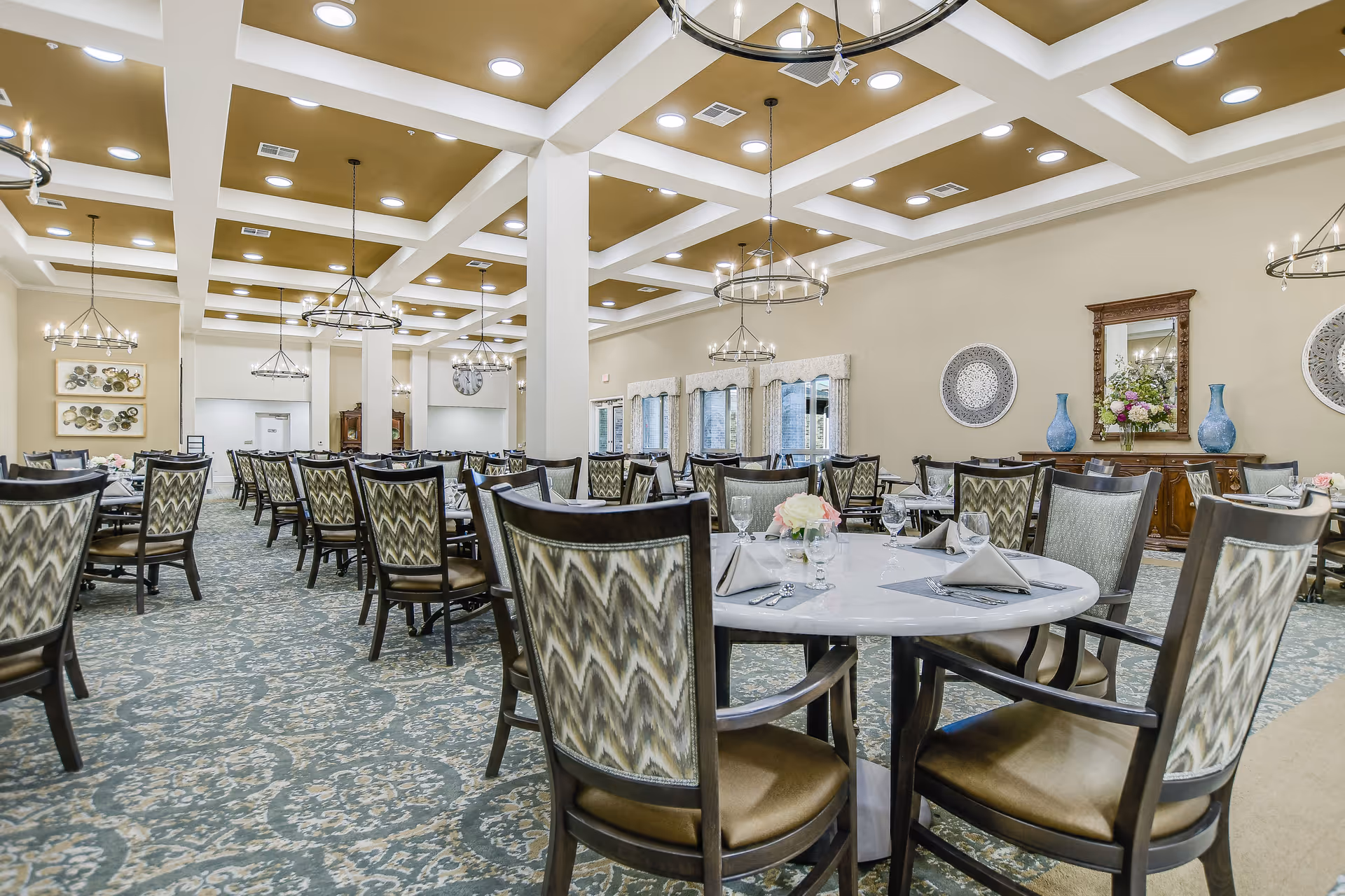 A spacious dining room with multiple round and rectangular tables set with napkins, glasses, and floral centerpieces. The room features patterned carpet, beige walls, large windows with curtains, decorative wall art, and modern chandeliers hanging from a coffered ceiling.