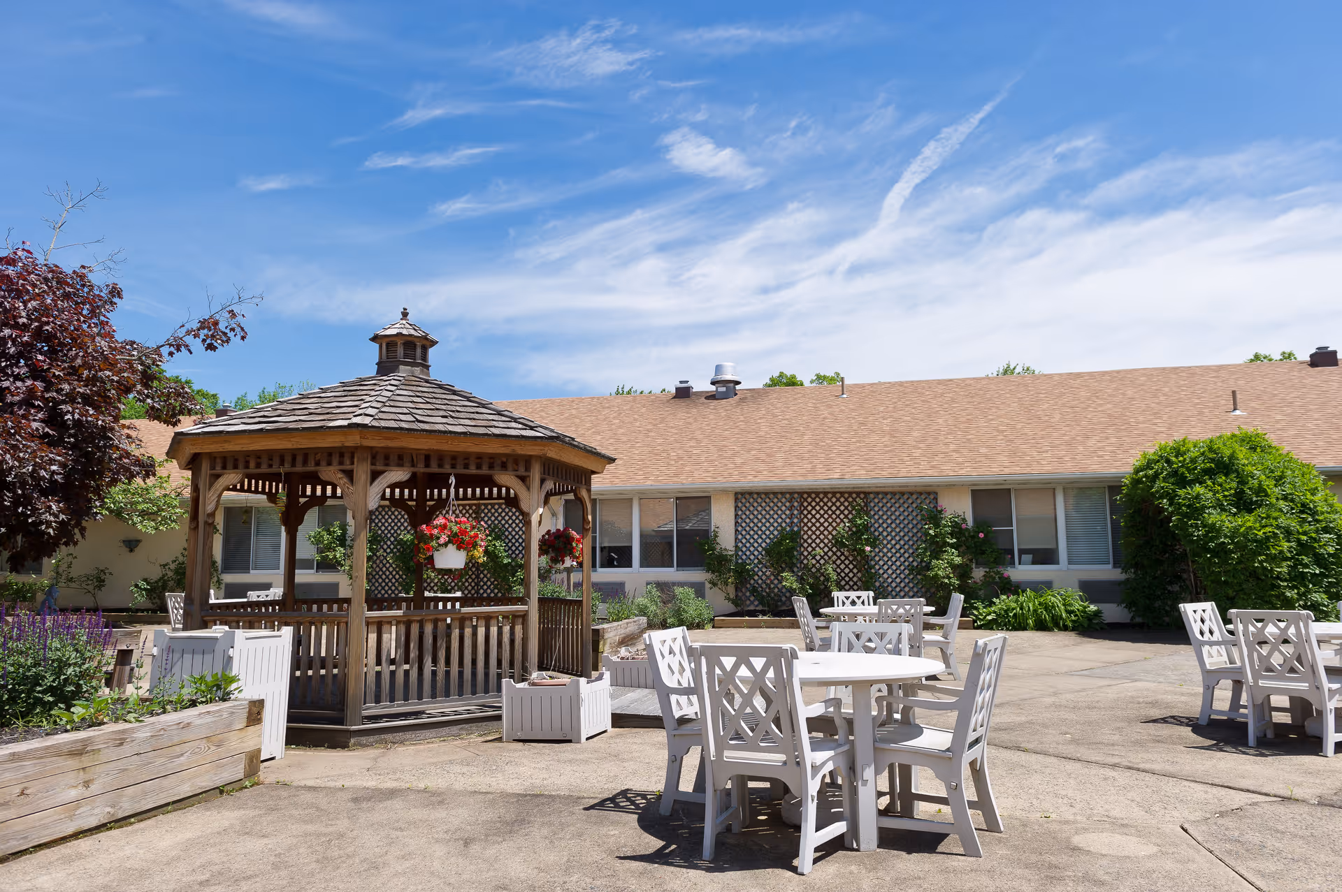 Outdoor patio area at Autumn Lake Healthcare at Berkeley Heights featuring a wooden gazebo with hanging flower baskets, several white tables and chairs arranged on a concrete surface, surrounded by greenery and a building with windows in the background under a blue sky with some clouds.