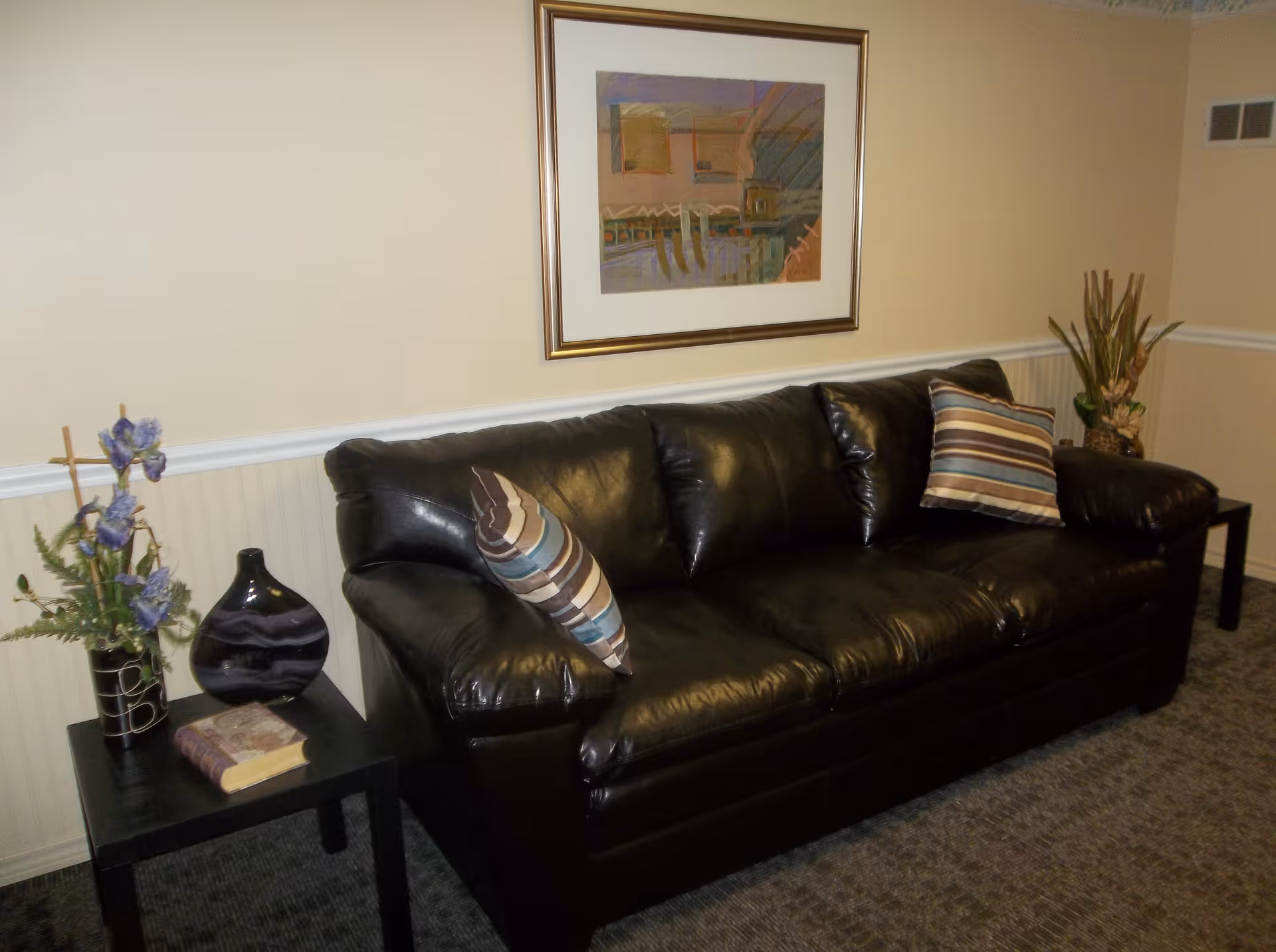 A black leather couch with two striped pillows in a room with beige walls and carpeted floor. There is a framed abstract painting hanging above the couch. On either side of the couch are small black tables, one with a vase containing purple flowers and a book, and the other with a decorative plant.