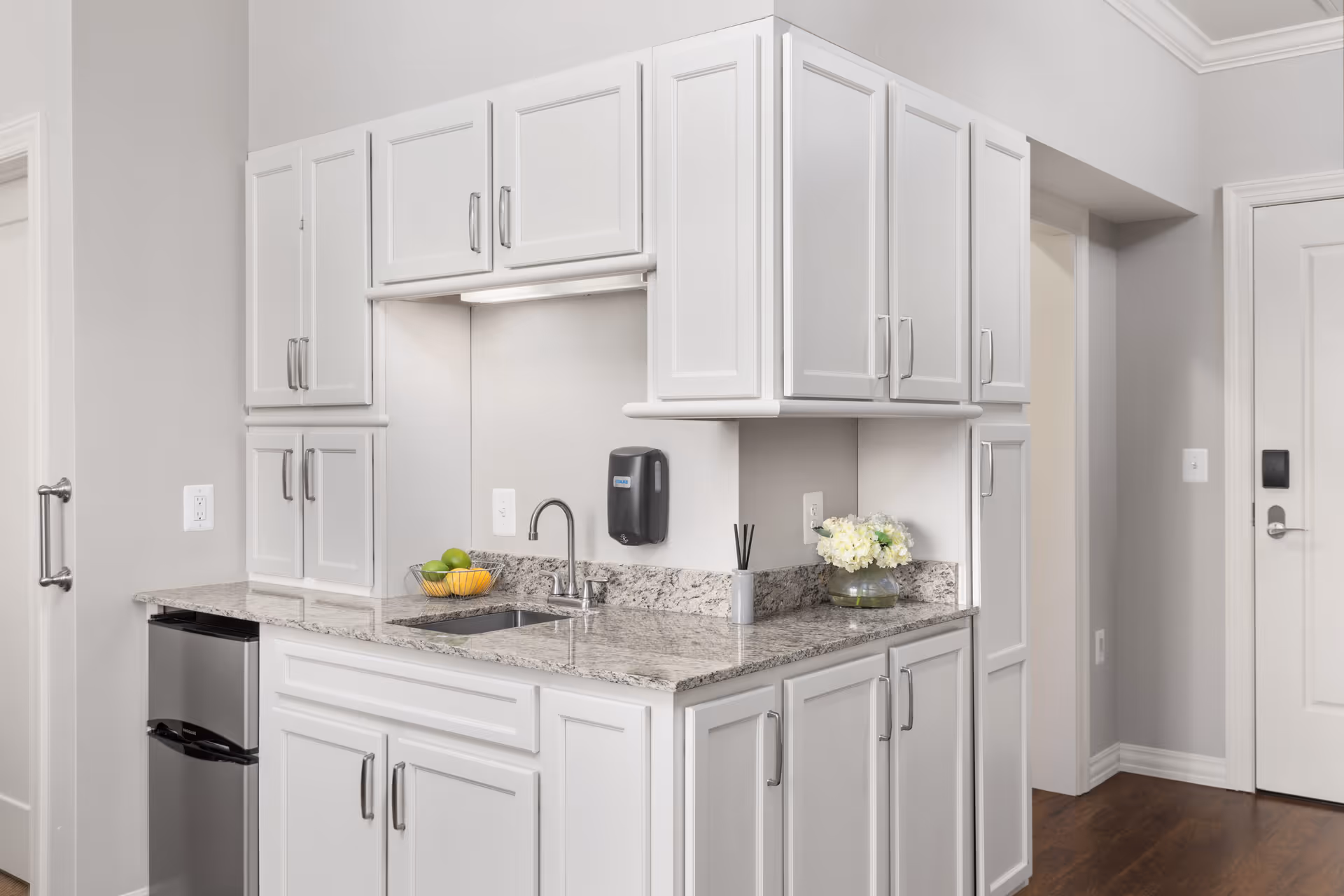 A clean, modern kitchenette with white cabinets, granite countertops, a small stainless steel refrigerator, a sink with a faucet, a basket of fruit, a vase with white flowers, and a wall-mounted soap dispenser. The floor is wooden, and there are white doors and light gray walls in the background.