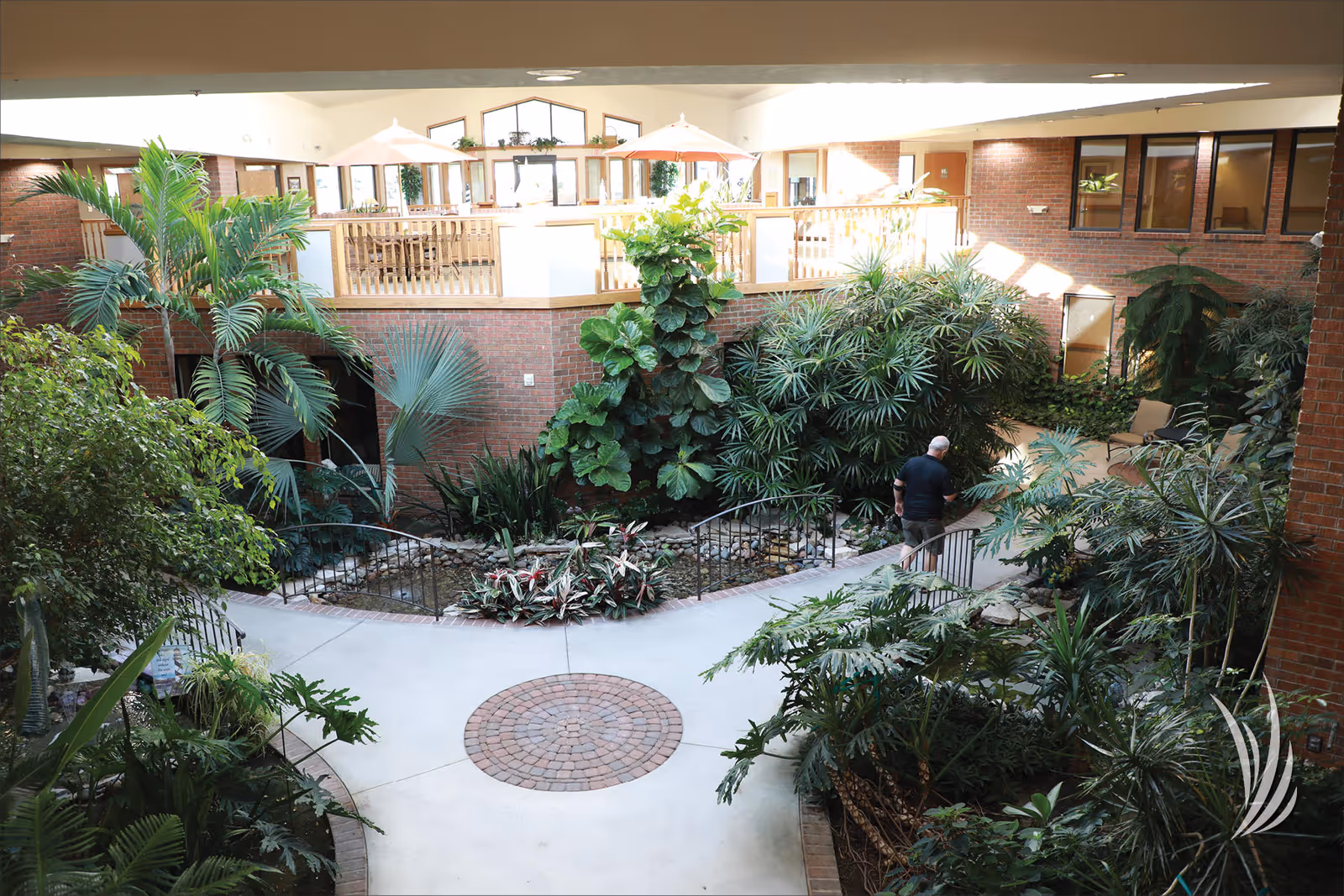 Indoor atrium with lush plants, a circular brick floor inset, walkways and a person walking beneath a second-floor balcony dining area.