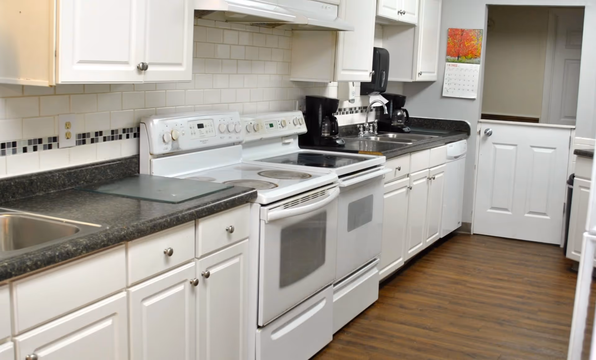 A clean, modern kitchen with white cabinets, black countertops, a white electric stove with oven, a double sink, two coffee makers, a dishwasher, and a wooden floor. A calendar with a colorful image is hanging on the wall next to a white door.