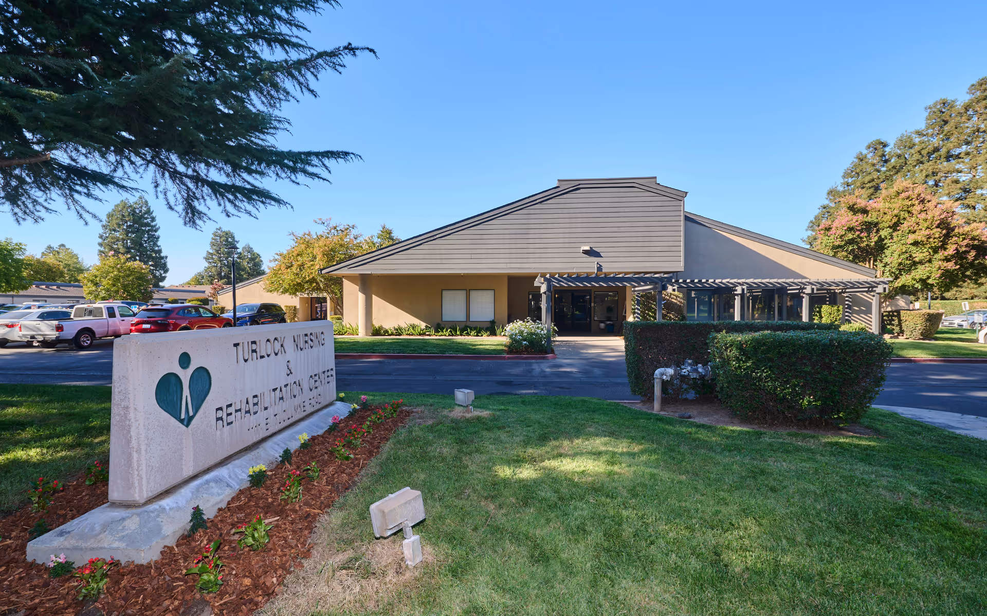 Front exterior of Turlock Nursing and Rehabilitation Center showing the entrance, landscaped lawn, and a stone sign in the foreground.