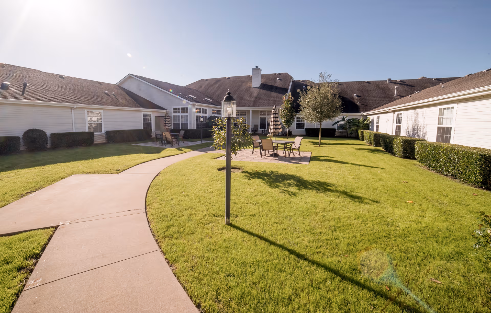Sunny courtyard with a winding sidewalk, green lawn, patio tables and chairs, and a surrounding single-story building.