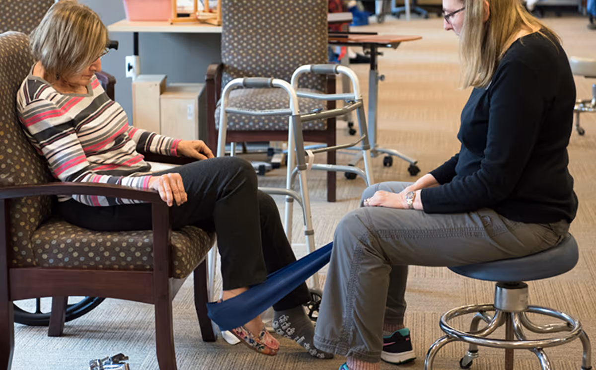 Two women in a senior living facility; one woman is seated in a chair with a resistance band around her feet, while the other woman, seated on a stool, assists her with a leg exercise. A walker and other chairs are visible in the background.