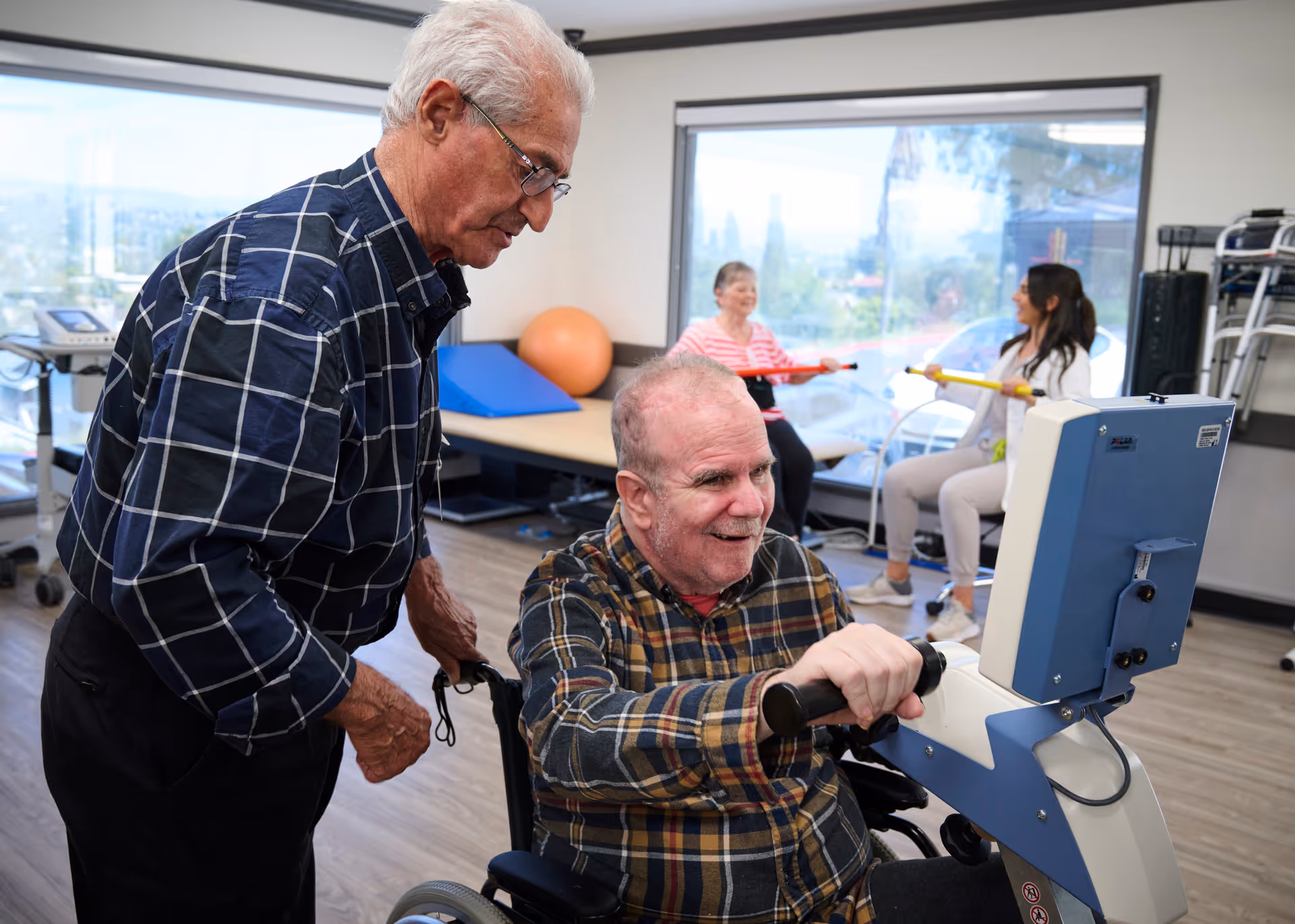 An elderly man in a wheelchair using an exercise machine with the assistance of another elderly man standing beside him. In the background, two women are seated and exercising with resistance bands in a bright room with large windows.