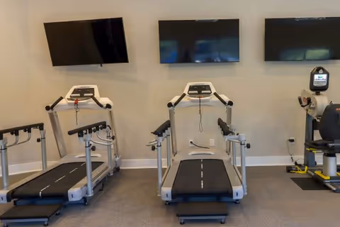 A small fitness room with treadmills and exercise equipment facing three wall-mounted flat-screen TVs.