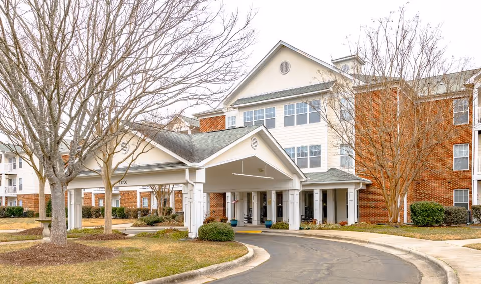 Exterior view of a multi-story senior living facility with a covered entrance driveway, brick and white siding facade, leafless trees, and a curved road leading to the entrance.