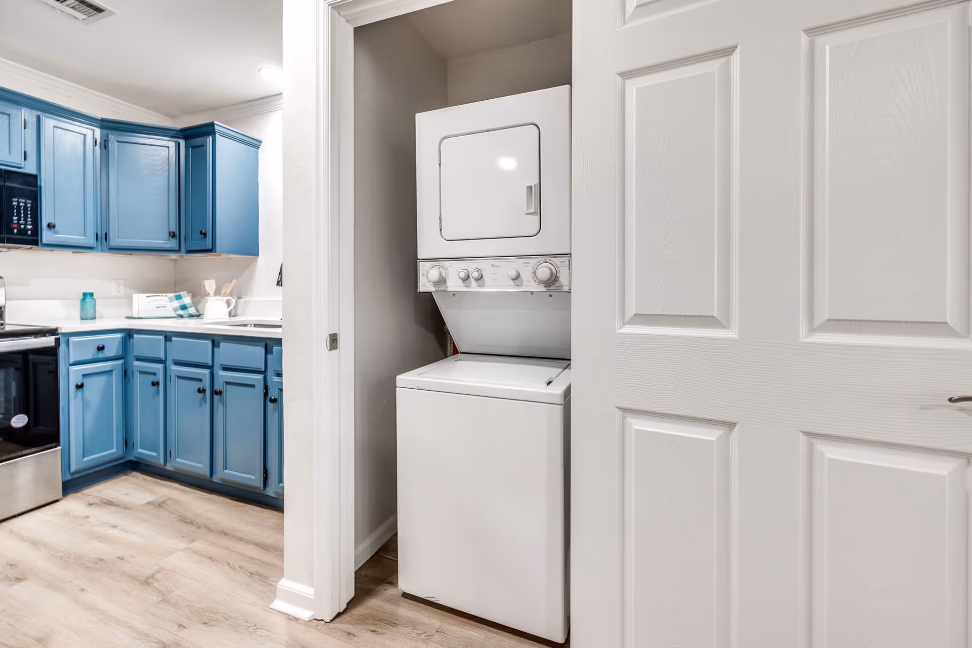 Interior view of a kitchen with blue cabinets and a stainless steel oven. To the right, there is a white stacked washer and dryer unit inside a small closet with a partially open white door.