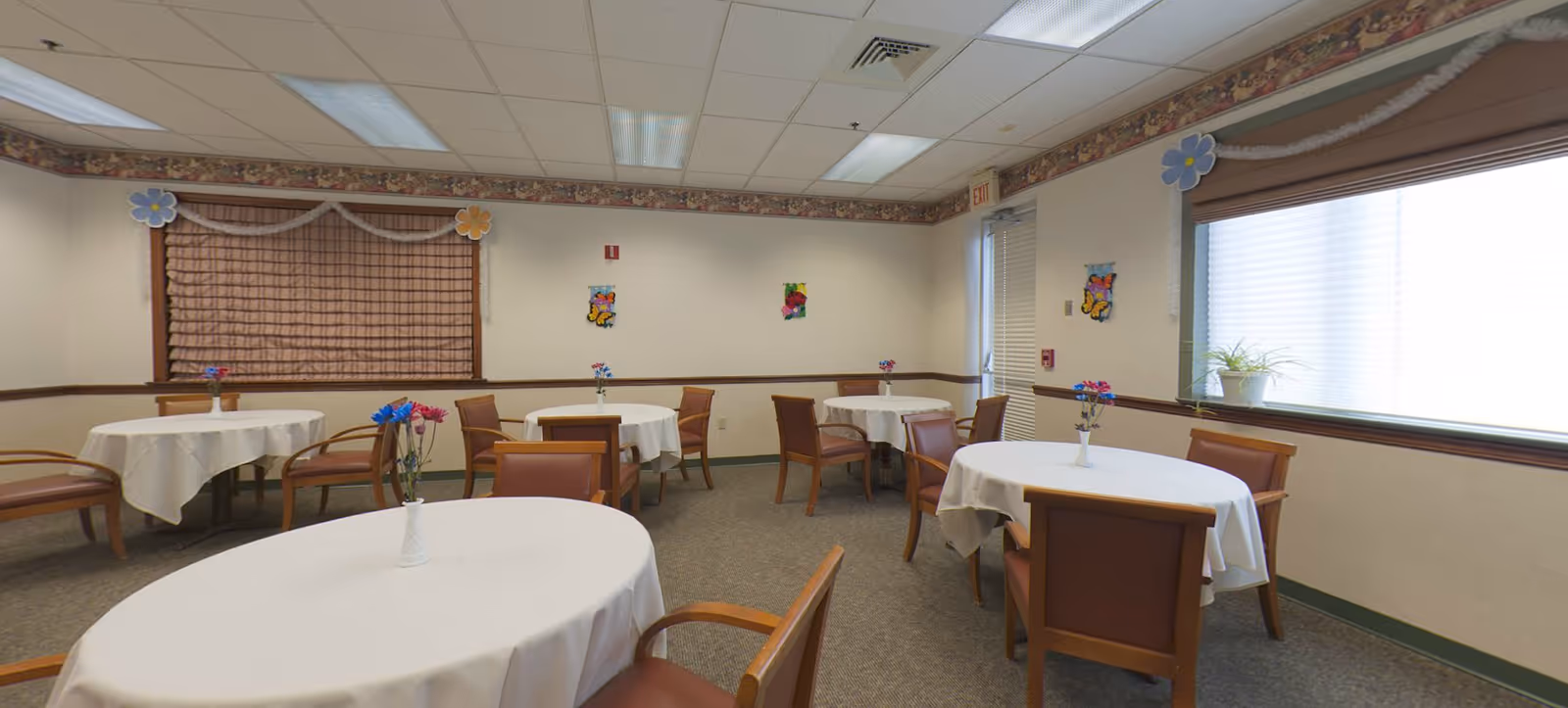 A dining room with several round tables covered with white tablecloths, each table having a small vase with colorful artificial flowers. The room has brown chairs with armrests, beige walls decorated with small butterfly and flower wall hangings, a window with blinds, and a ceiling with fluorescent lights.