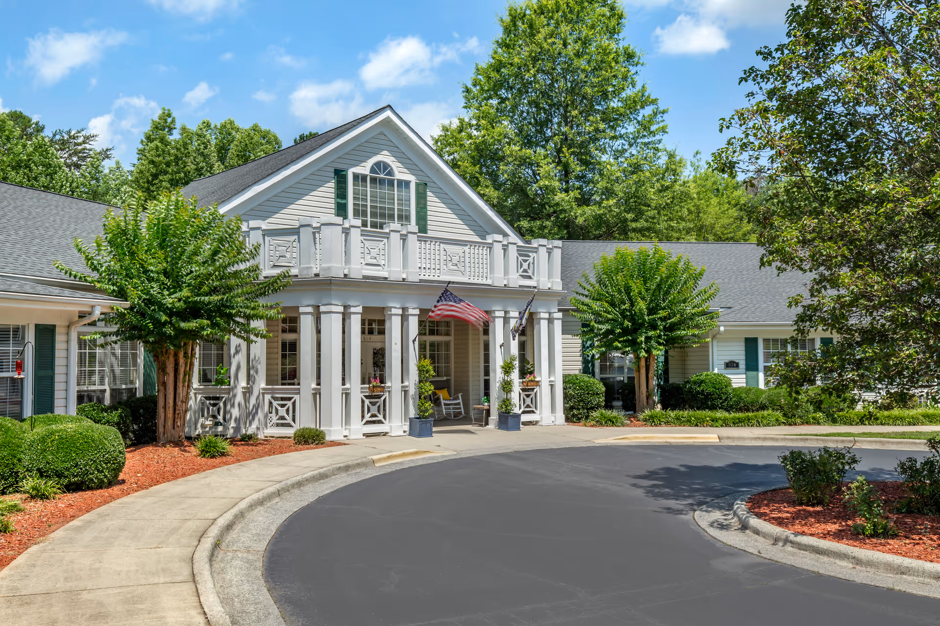 Front exterior view of a senior living facility building with white columns, a balcony, and two flags displayed near the entrance. The building is surrounded by green trees, bushes, and landscaped areas with red mulch under a blue sky with some clouds.