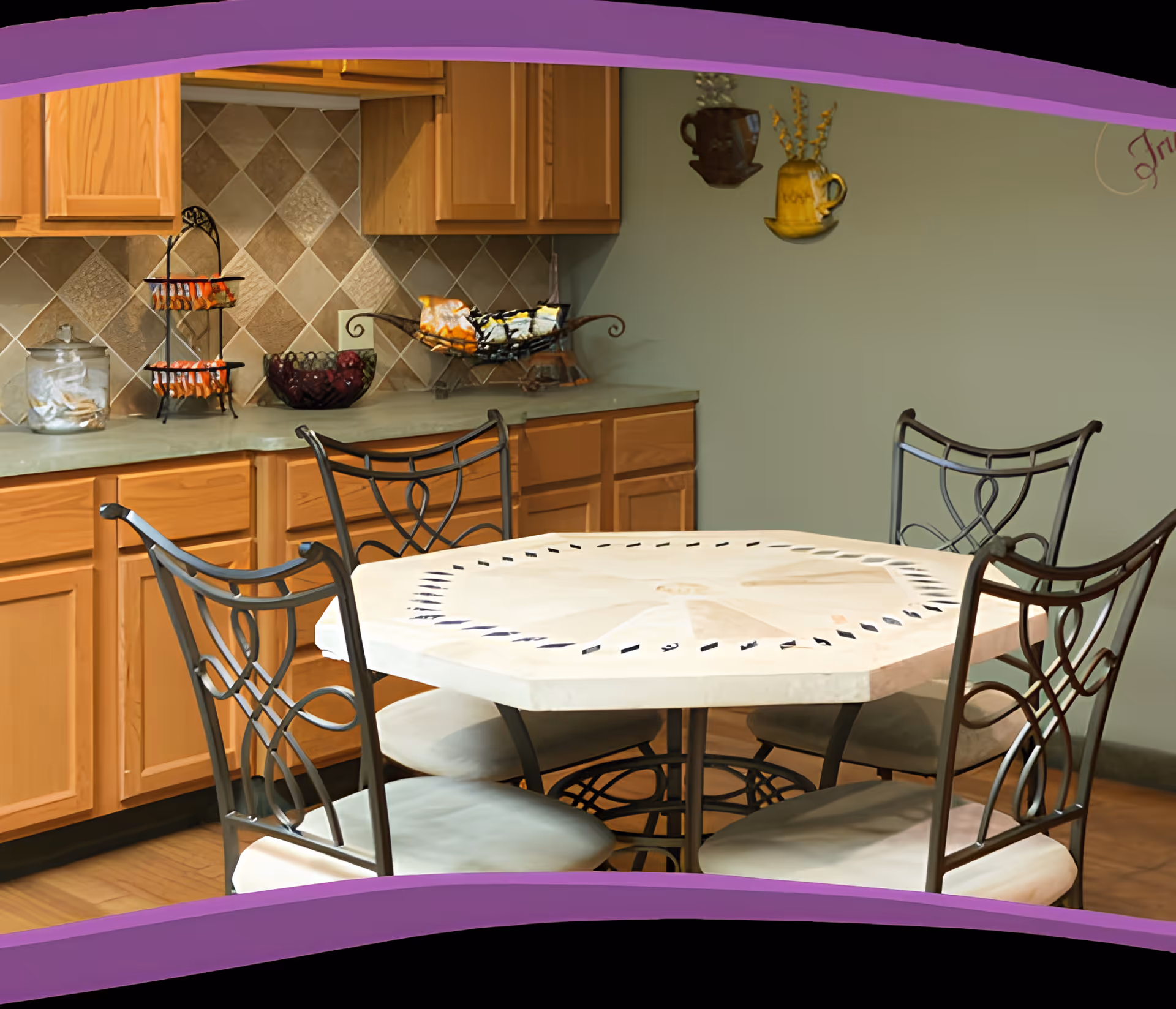 A kitchen area with wooden cabinets and a tiled backsplash. In the foreground, there is a round dining table with a decorative white top and four metal chairs with cushioned seats. On the countertop, there are containers with snacks and a decorative bowl. The wall is painted green with two decorative hanging mugs.
