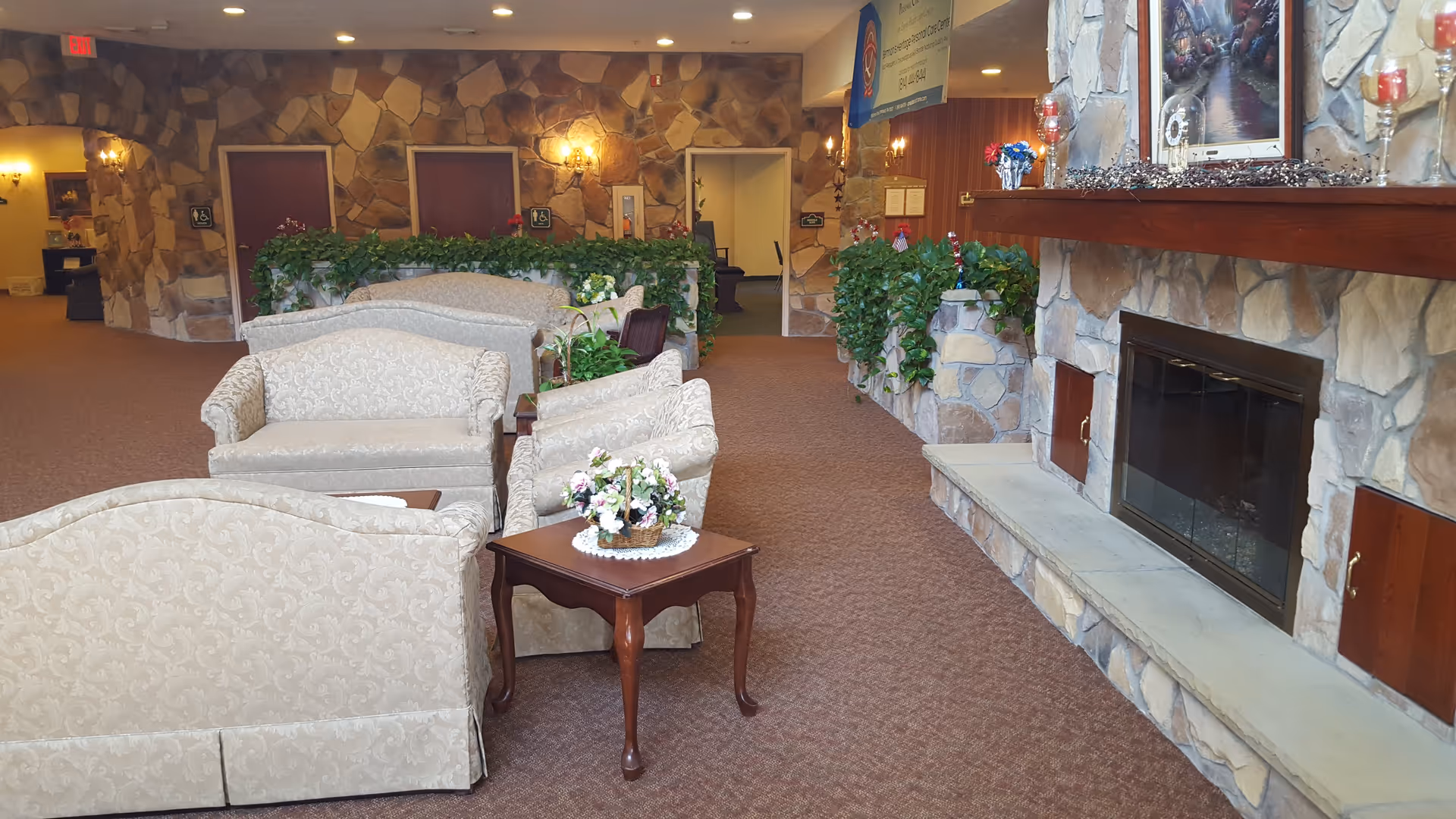 A cozy lounge area in Siemon's Heritage Personal Care Center featuring beige patterned sofas and armchairs arranged around small wooden tables with floral decorations. The room has stone walls and a large stone fireplace with a wooden mantle adorned with candles and a framed painting. There are green plants along the stone half-walls and warm lighting from wall sconces. In the background, there are doors and an open hallway.