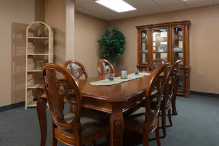 A dining room with a long wooden table surrounded by eight ornate wooden chairs. On the table is a light green runner with decorative candle holders. In the background, there is a wooden china cabinet with glass doors displaying dishes, a tall green potted plant, and a beige shelving unit with some items on it. The room has beige walls and a carpeted floor.