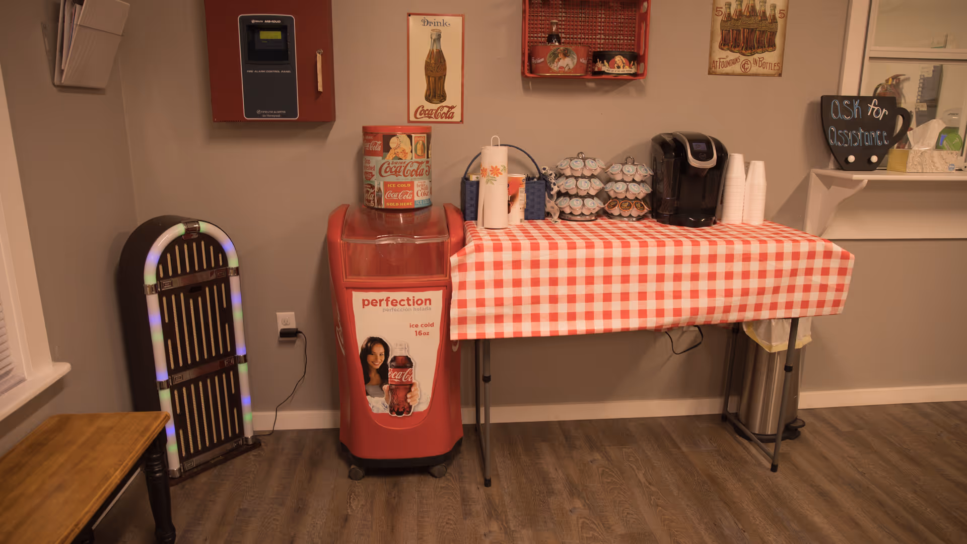 A refreshment area with a red and white checkered tablecloth on a folding table holding a coffee maker, disposable cups, paper towels, and coffee pods. Next to the table is a vintage-style Coca-Cola cooler with a Coca-Cola sign above it. A small jukebox is placed against the wall to the left, and a chalkboard sign on a shelf to the right reads 'Ask for Assistance'. The floor is wood, and the walls are painted light gray.