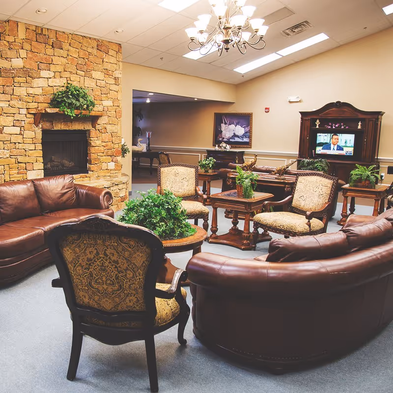 A cozy senior living facility lounge area with brown leather sofas and upholstered chairs arranged around wooden tables with green plants. A stone fireplace is on the left wall, and a wooden cabinet with a television is on the right. The room has beige walls and a chandelier hanging from the ceiling.