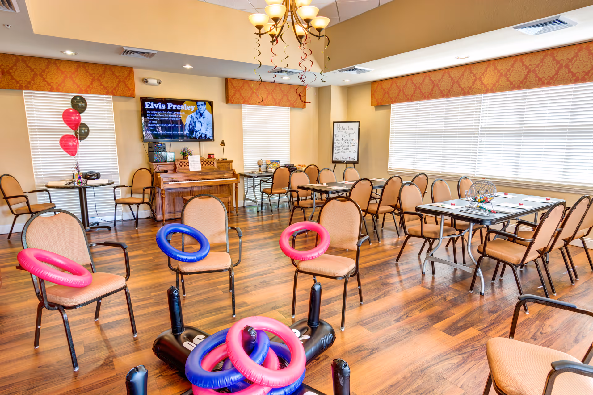 Bright communal activity room with chairs and tables, a piano and TV displaying Elvis Presley, and colorful inflatable rings for games.