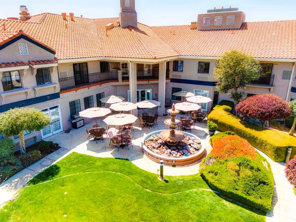 Outdoor courtyard area of a senior living facility with a central stone fountain surrounded by patio tables and chairs with umbrellas. The courtyard is bordered by well-maintained green lawns, trimmed bushes, and trees. The building has a tiled roof and balconies overlooking the courtyard.