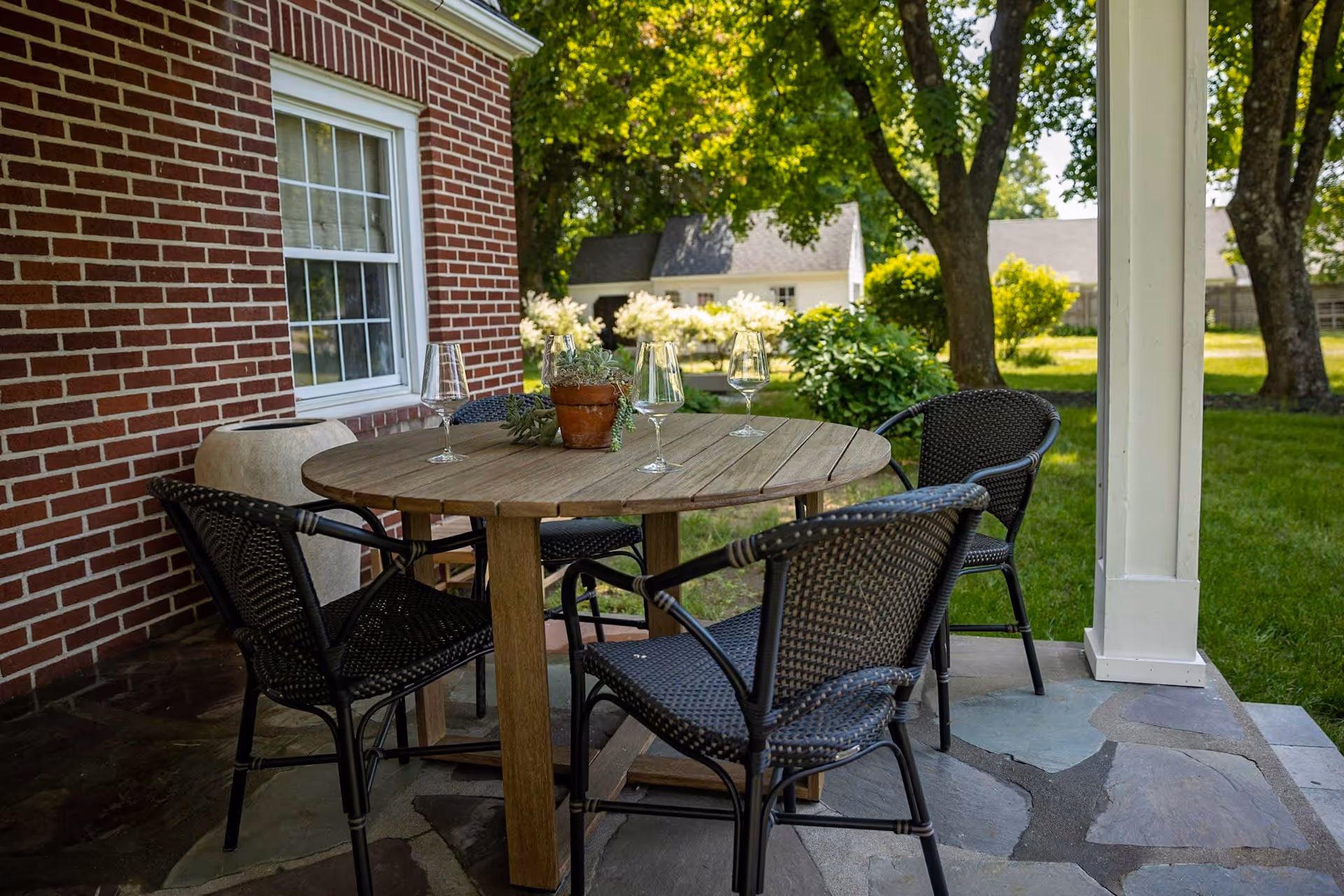 Outdoor patio area with a round wooden table surrounded by four black wicker chairs. On the table are four empty wine glasses and a small potted plant. The patio is next to a red brick building with a window, and the background shows green grass, trees, and a white house under a sunny sky.
