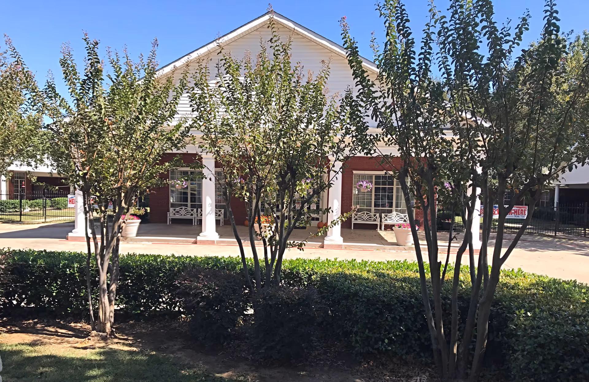 Front exterior view of a single-story building with white columns and a triangular roof, partially obscured by several small trees and bushes in the foreground. There are benches and flower pots on the porch area under the roof.