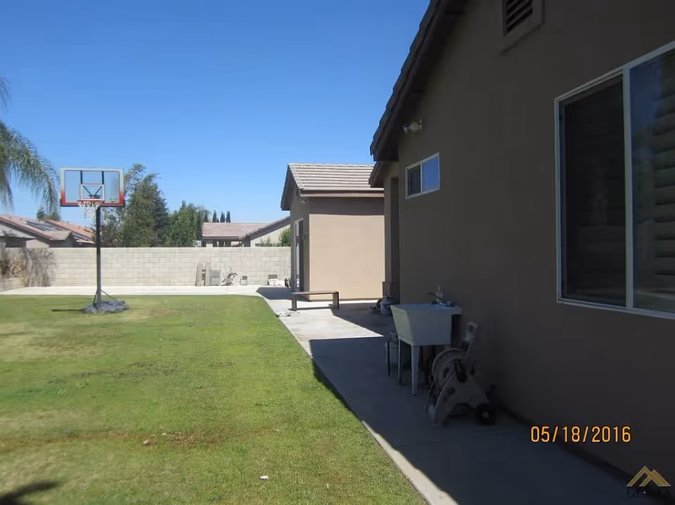Outdoor view of a backyard area with a basketball hoop on a grassy lawn, a concrete pathway alongside a beige house, and a small utility sink with a hose reel attached to the house wall under a clear blue sky.