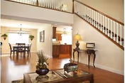 Interior view of a senior living facility showing a spacious area with a wooden staircase on the right, a small table with a lamp and decorative items underneath the stairs, a dining area with a table and chairs in the background, and a kitchen area beyond the dining space. The floor is wooden, and the space is well-lit with natural light coming through a window.