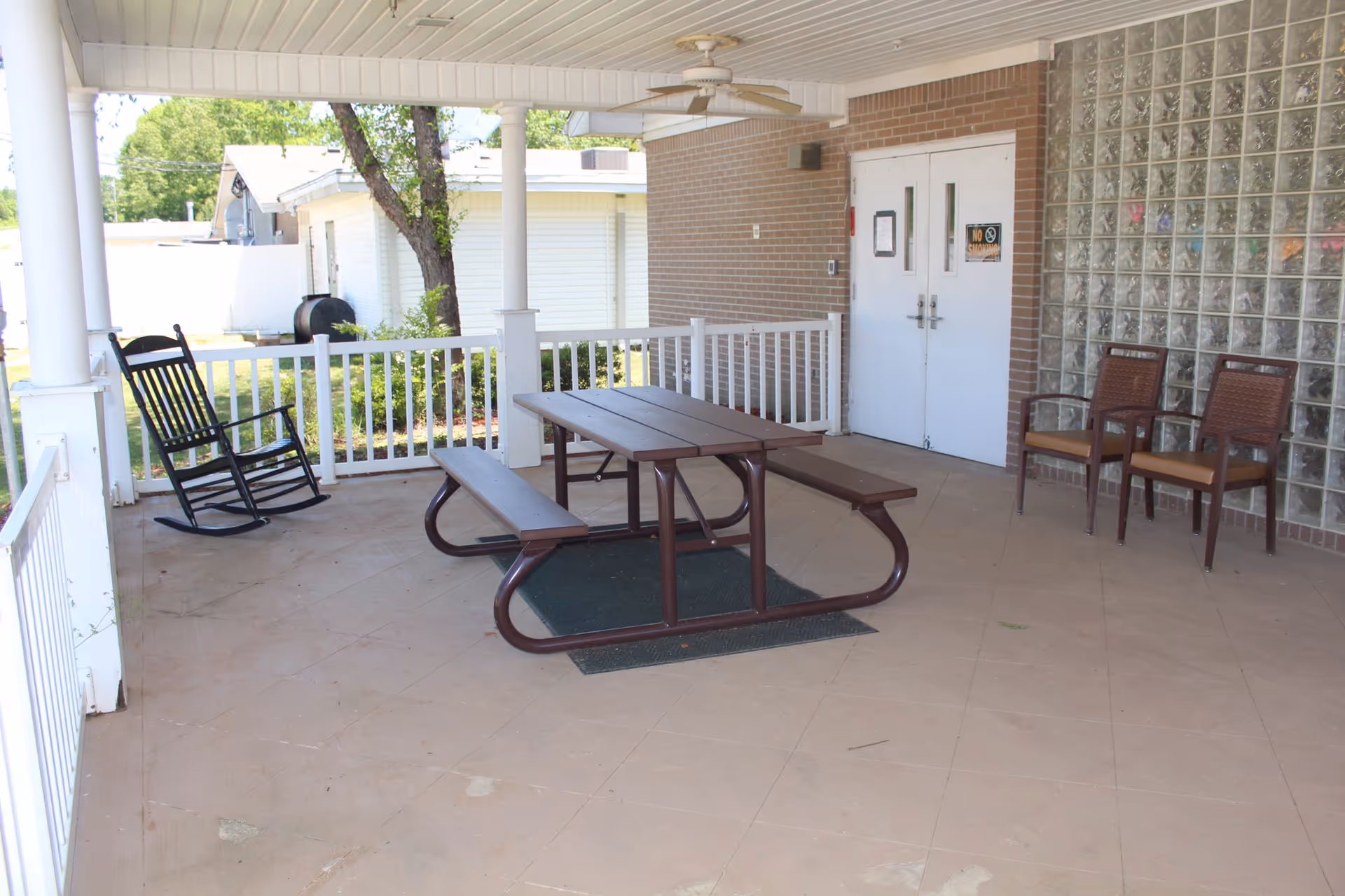 Covered outdoor patio area with a brown picnic table and benches, two brown chairs with armrests, a black rocking chair, white railing, ceiling fan, and double white doors leading inside a building.