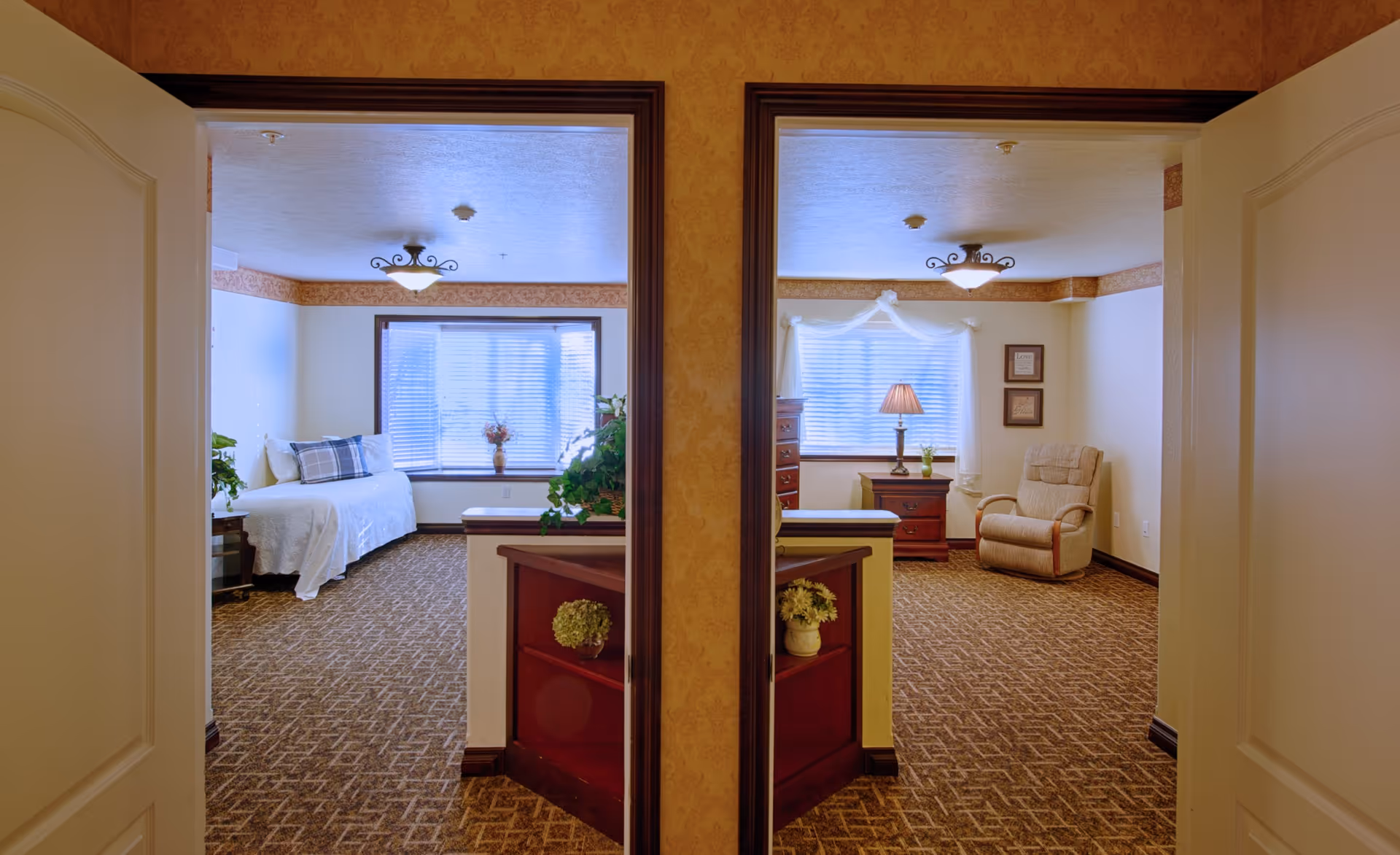 View through two open doors into two adjacent rooms in a senior living facility. The left room features a single bed with white bedding and a plaid pillow, a side table with a plant, and a large window with blinds. The right room has a comfortable armchair, a wooden dresser, a side table with a lamp and a small plant, framed wall art, and a window with sheer curtains and blinds. Both rooms have patterned carpet and warm lighting.