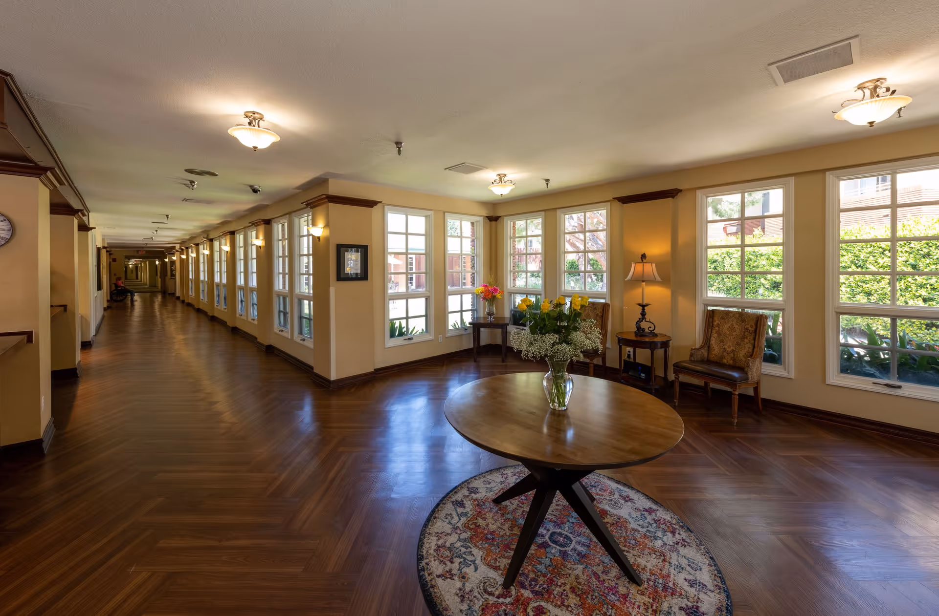 A spacious, well-lit hallway in a retirement community with large windows along one side letting in natural light. The hallway features wooden flooring and beige walls with decorative molding. At the near end, there is a round wooden table with a vase of flowers on a colorful rug, flanked by two upholstered chairs and a small side table with a lamp. The far end of the hallway shows a person in a wheelchair.