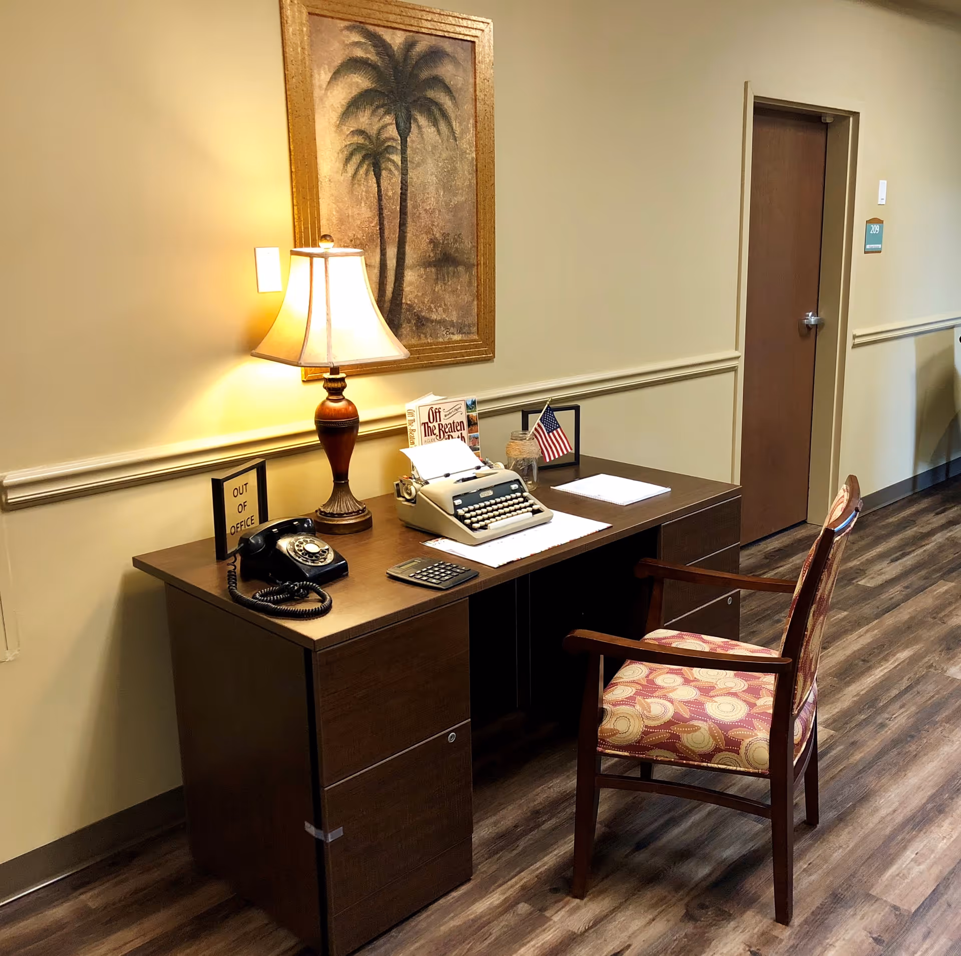 A wooden desk with a vintage rotary phone, a typewriter, a calculator, a lamp, and small framed signs including one that says 'OUT OF OFFICE' and a small American flag. The desk is placed against a beige wall with a framed painting of palm trees above it. A patterned wooden chair is positioned in front of the desk, and a closed door labeled 209 is visible in the background.