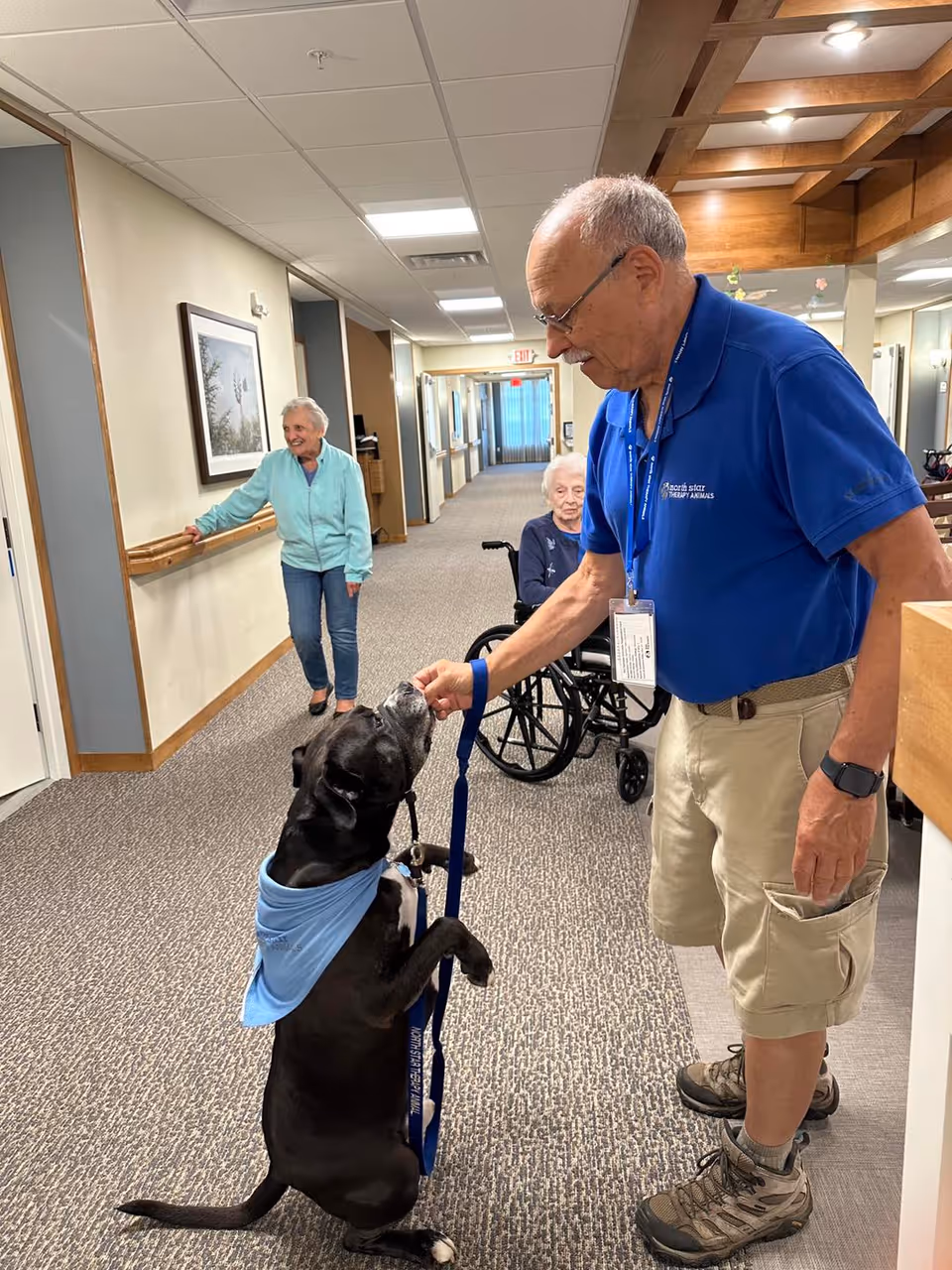 An elderly man in a blue shirt and khaki shorts is feeding a black dog wearing a blue bandana in a hallway of a senior living facility. In the background, an elderly woman in a wheelchair and another elderly woman standing near the wall are watching the interaction. The hallway has carpeted floors, beige walls, and framed artwork.